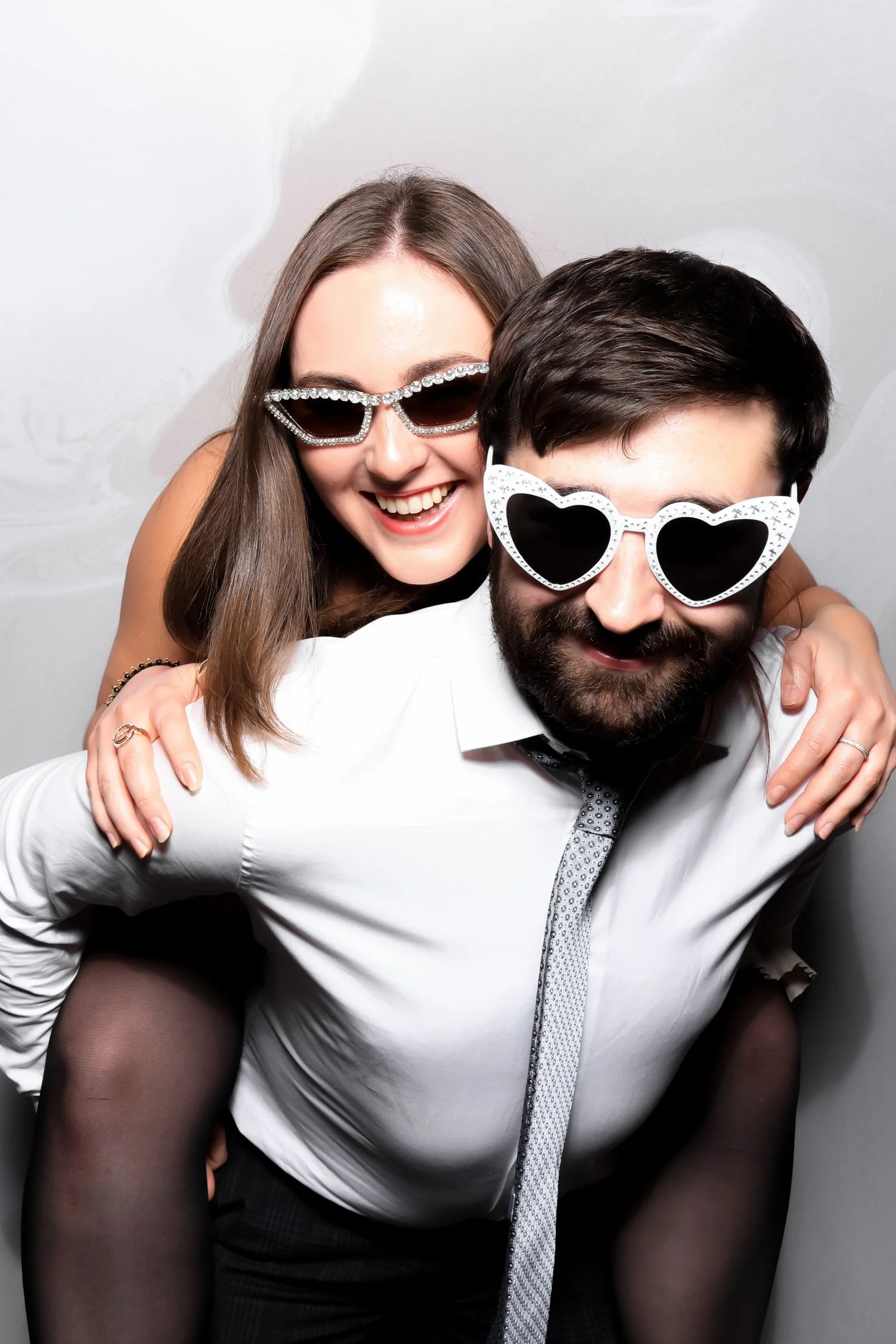 A smiling woman and a man wearing large sunglasses, with the woman riding on the man's back, both having fun.