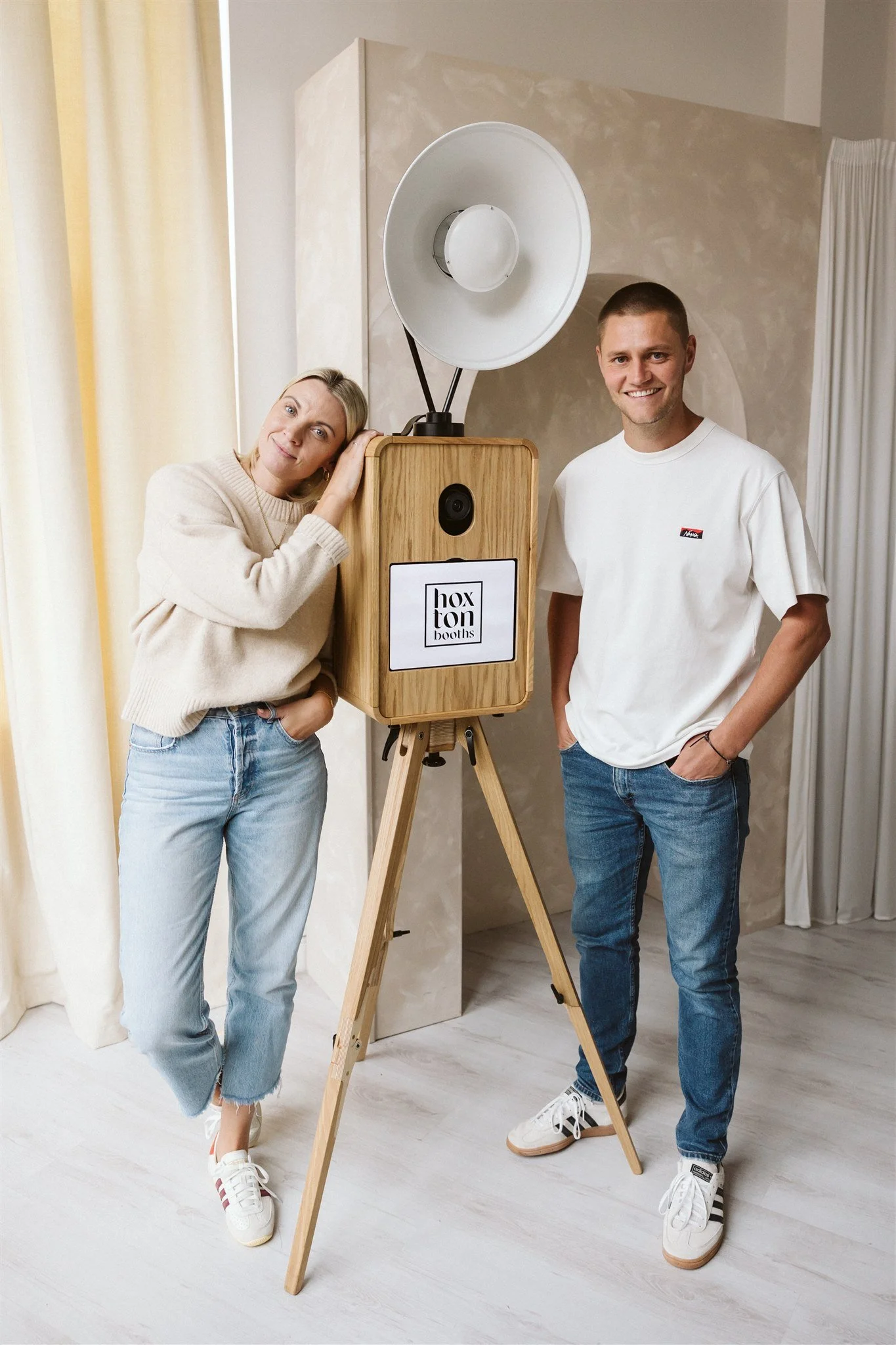 Two people standing next to a wooden photo booth with a sign that says "hoxton booths." One person is a woman leaning her head on the photo booth and the other is a man standing with his hands in his pockets, both smiling.