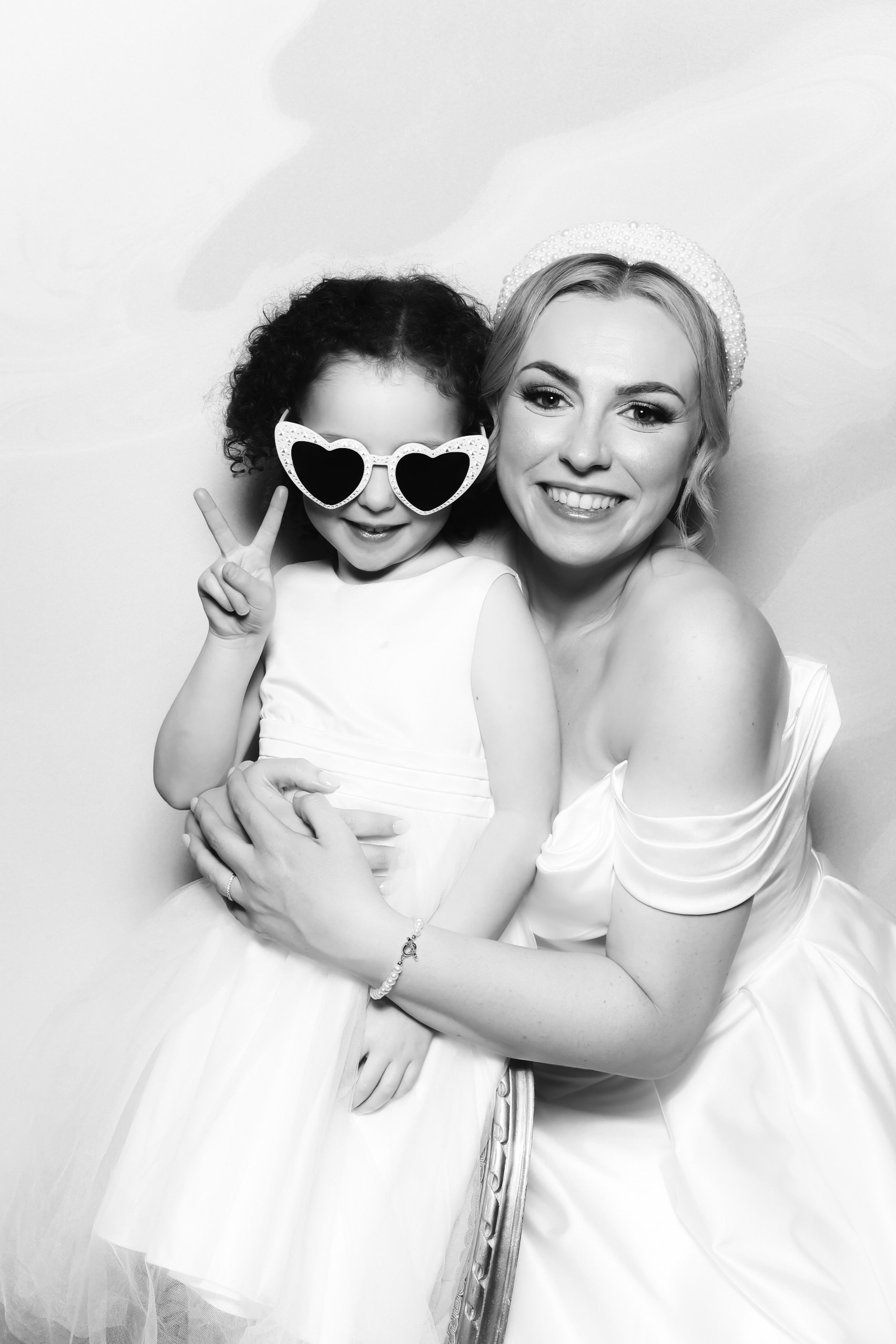 A woman and a young girl smiling together, with the girl making a peace sign and wearing heart-shaped sunglasses, both dressed in white, in a black-and-white photo.
