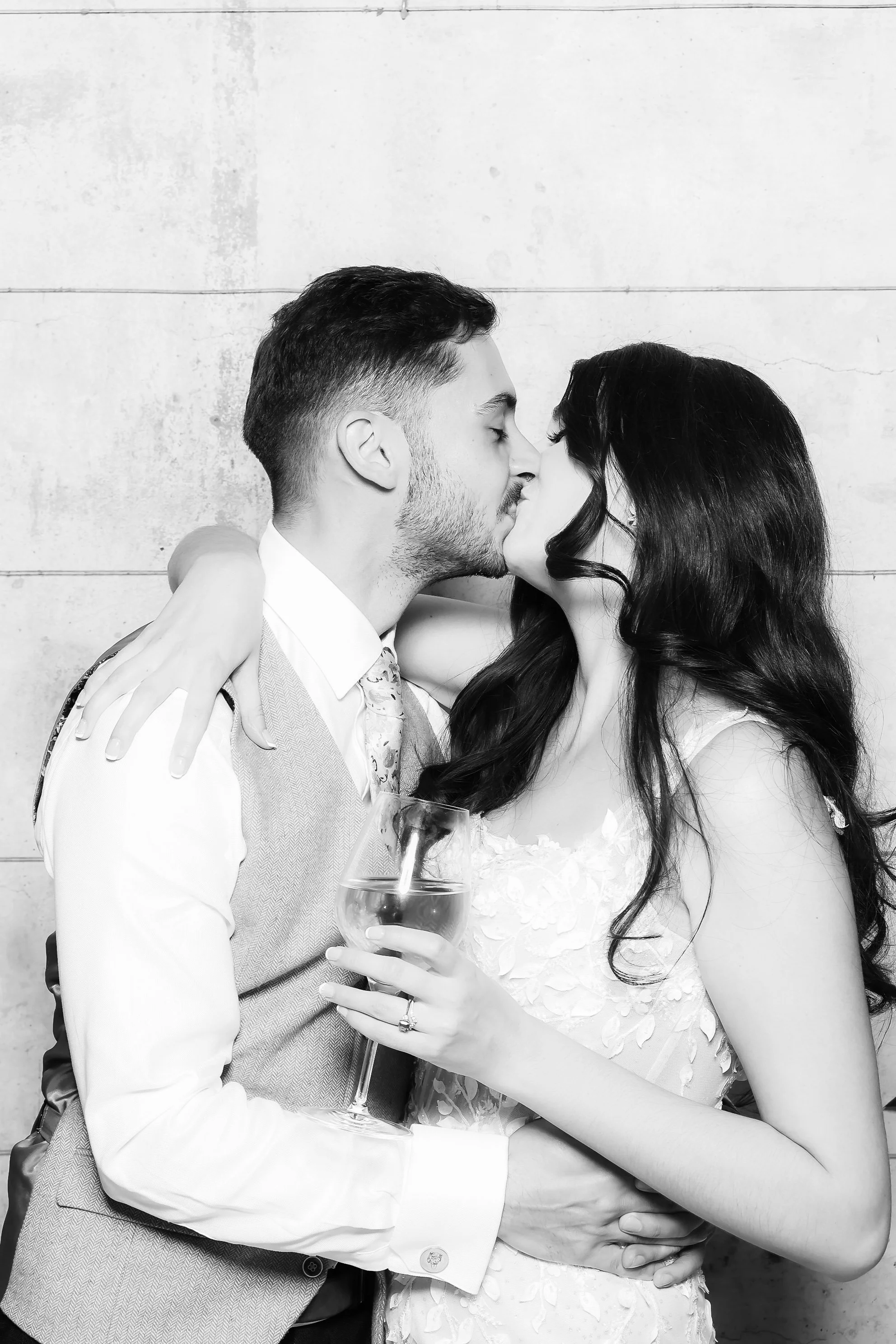 Black and white photo of a couple kissing, the man is holding a glass of wine, she has her arm around him, both are dressed in formal attire, standing against a plain wall.
