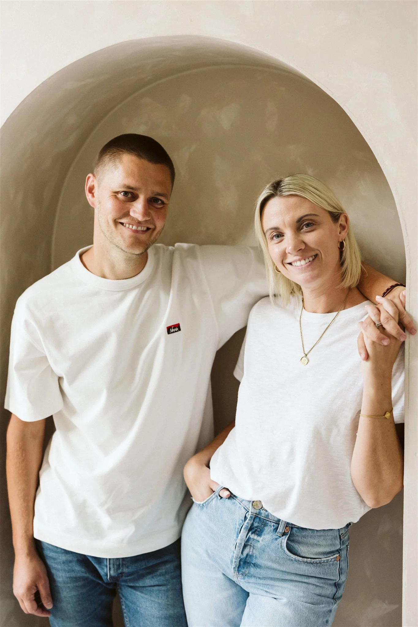 A man and woman smiling and posing together inside a small arched niche. They are both dressed casually in white T-shirts and light jeans, with the woman wearing gold jewelry and the man holding her shoulder.