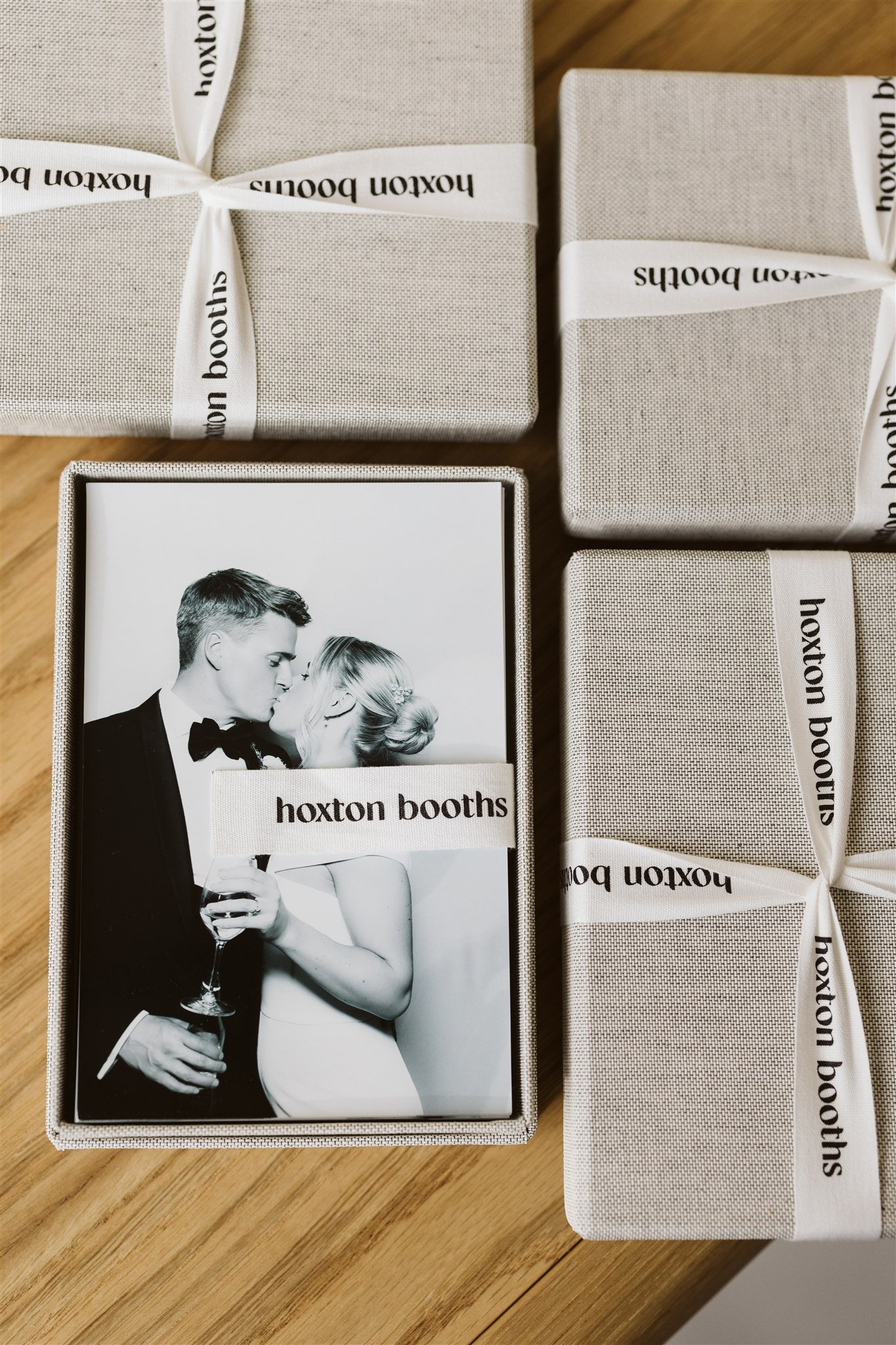 A black and white photograph of a couple kissing, with the woman holding a glass of wine. The photo is inside a box with a ribbon that says 'hoxton booths', surrounded by four wrapped gift boxes with similar ribbons.