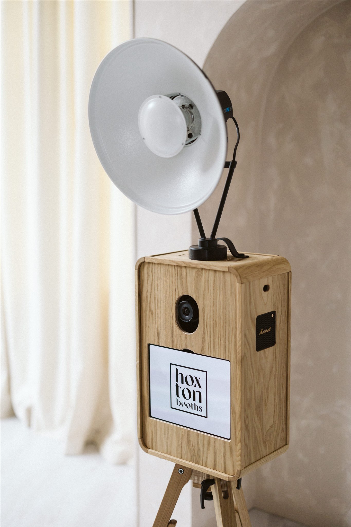 A wooden photo booth with a camera and a large LED light on top, standing on tripod legs, with a sign that reads 'hoxton booths' on the front.