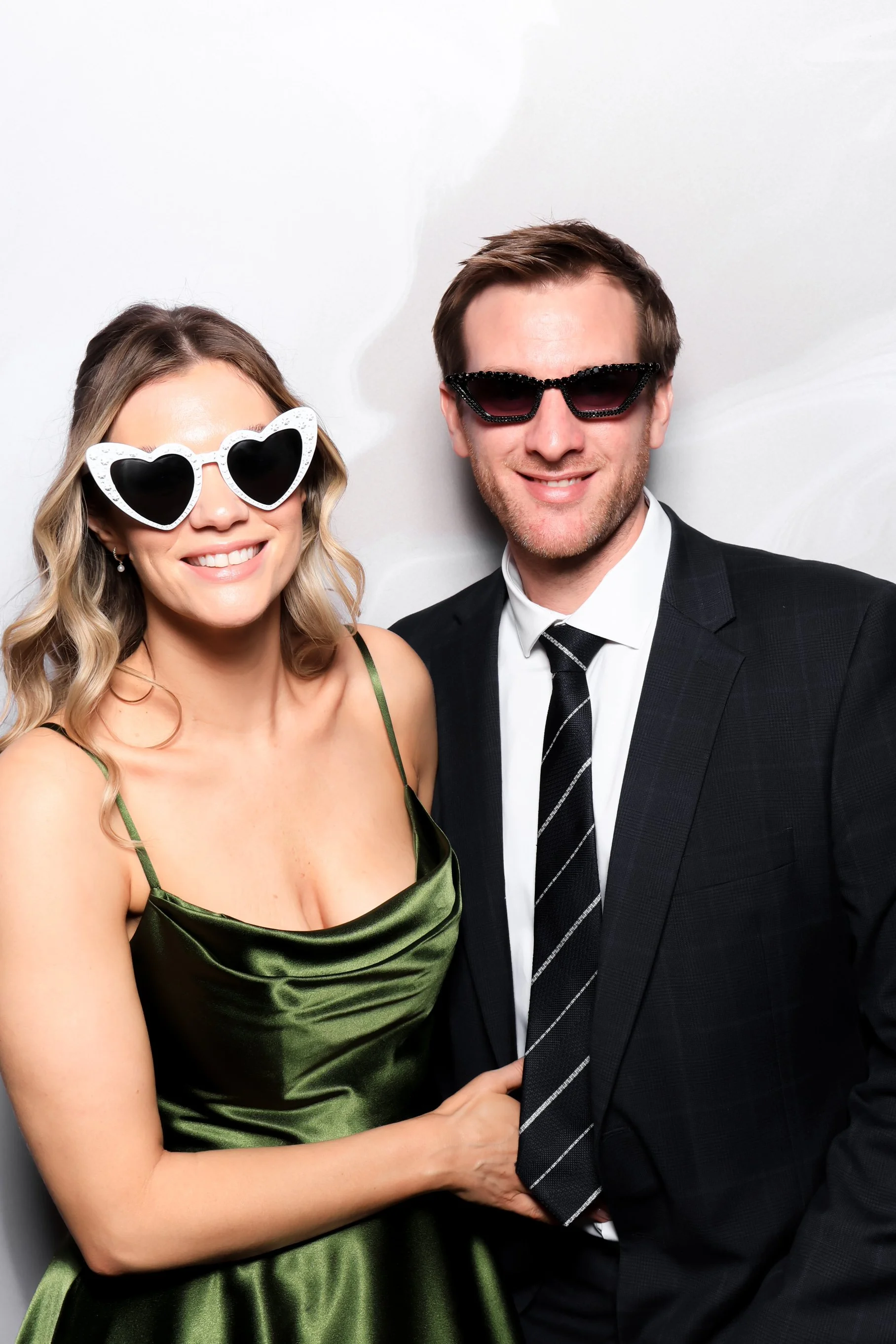 A man and woman dressed in formal attire, both wearing sunglasses, smiling and posing together.