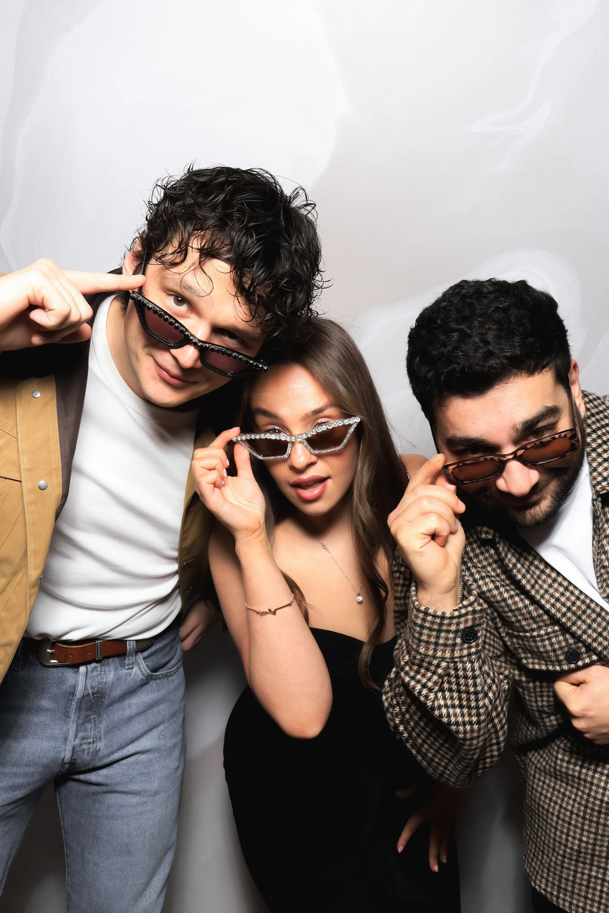 Three young people dressed stylishly, posing for a photo with playful expressions and sunglasses, against a plain background.
