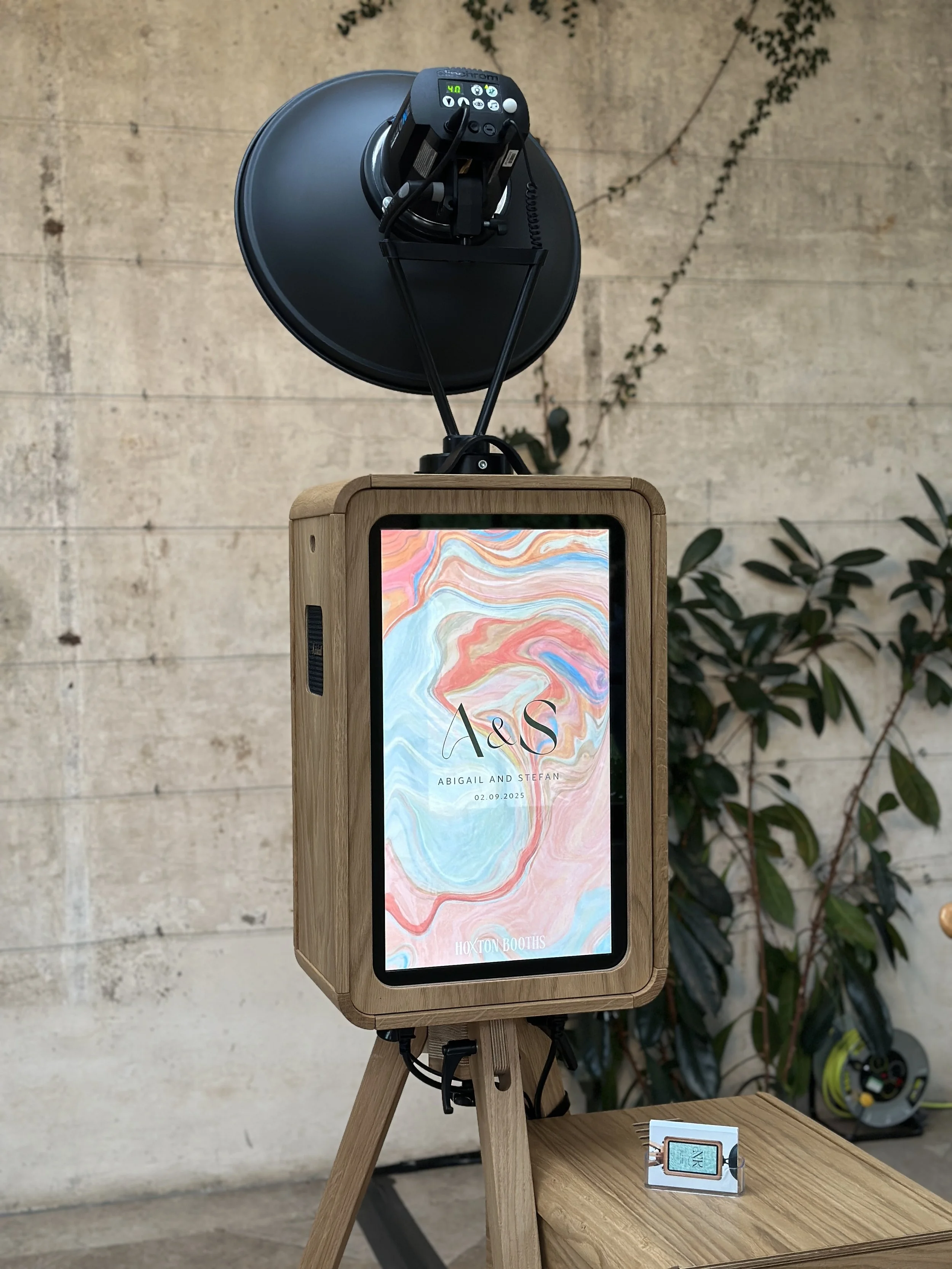 A photo booth with a digital screen floral artwork and text, set on a wooden tripod stand with a large mounted on top. There is a small sign with a QR code on the table in front of it, a textured wall in the background, and some plants nearby.