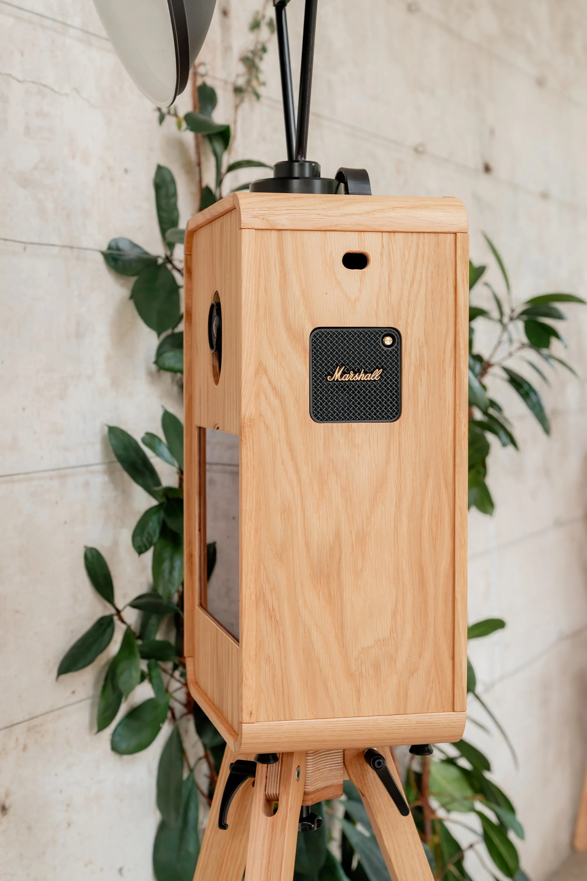 A luxury vintage-style wooden photo booth speaker standing on a wooden tripod, with a leafy plant in the background.