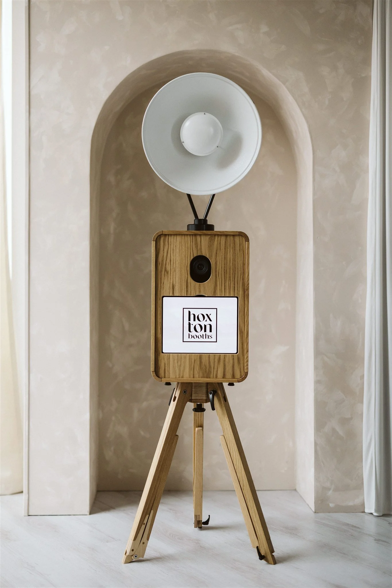 Photo of a vintage wooden photo booth on a tripod stand with a camera, a lamp on top, and the logo "Hoxton Booths" on the front.