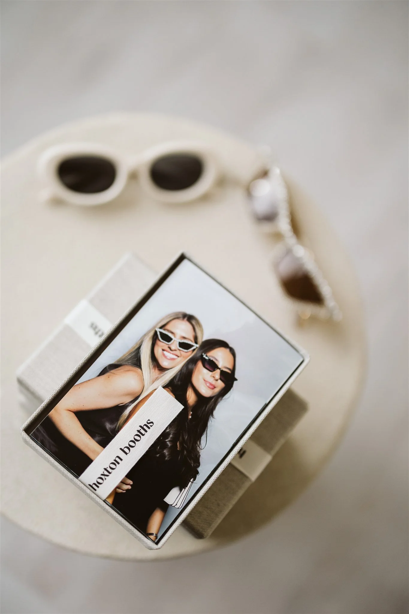 A photo booth picture of two women wearing sunglasses and smiling, with a card labeled 'Hoxton Booths', placed on a white surface along with a pair of white sunglasses and silver jewelry.
