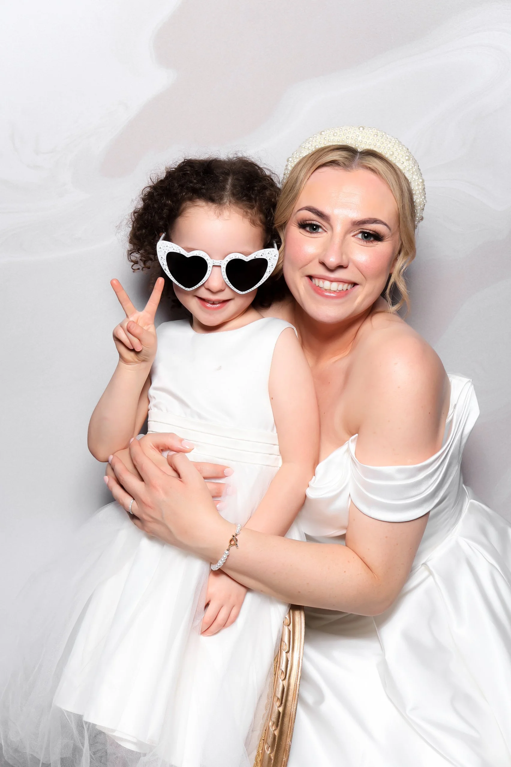 A smiling woman and young girl in white dresses posing together, with the girl wearing heart-shaped sunglasses and making a peace sign.