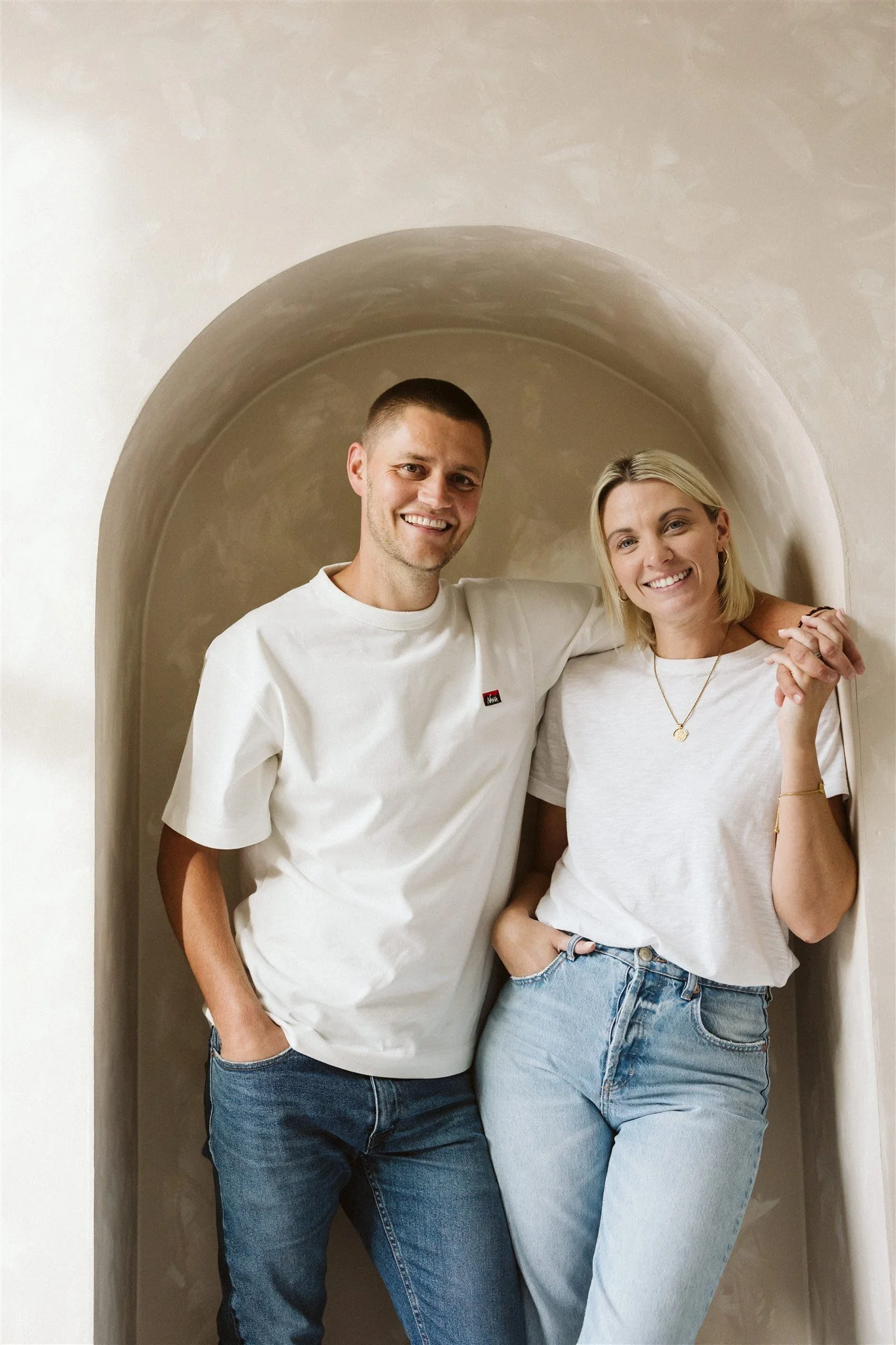 A smiling man and woman standing together in front of a rounded wall niche, with the man's arm around the woman's shoulder, both wearing casual white t-shirts and blue jeans.