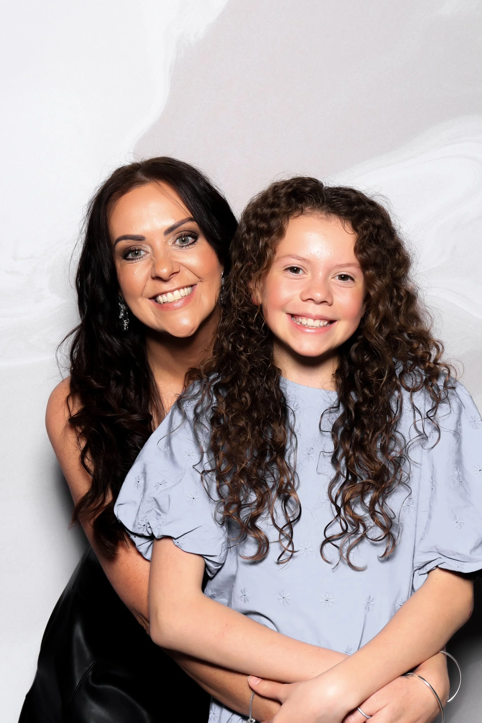 A woman and a young girl smiling and posing together against a light gray background.