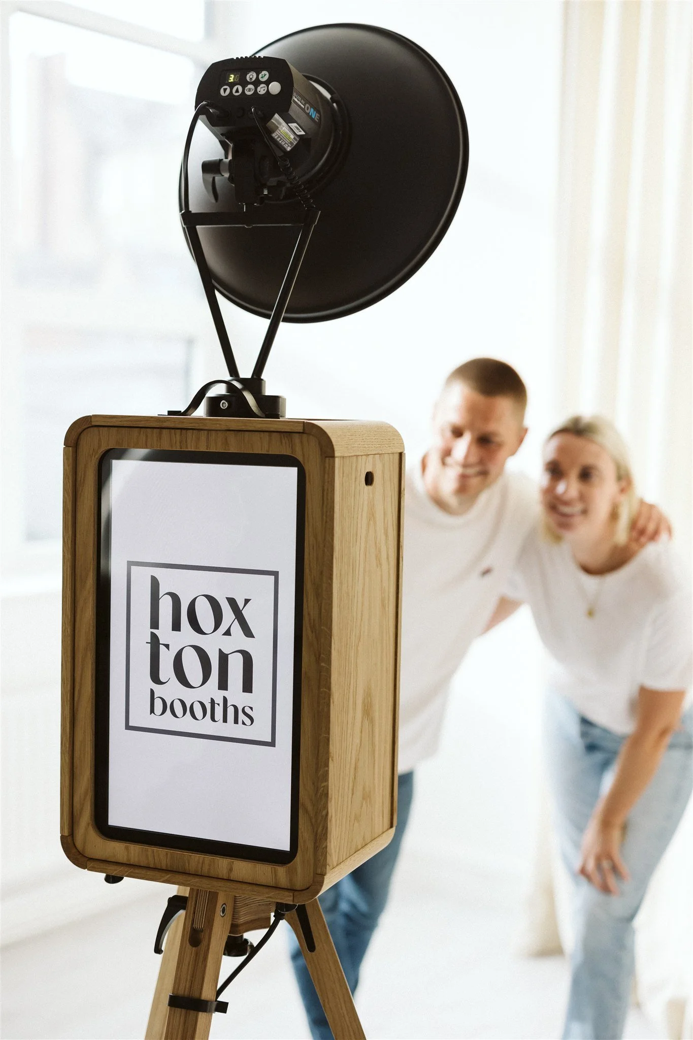 A photo of a photo booth with the word 'hoxton booths' on the screen, and a smiling couple posing in the background.