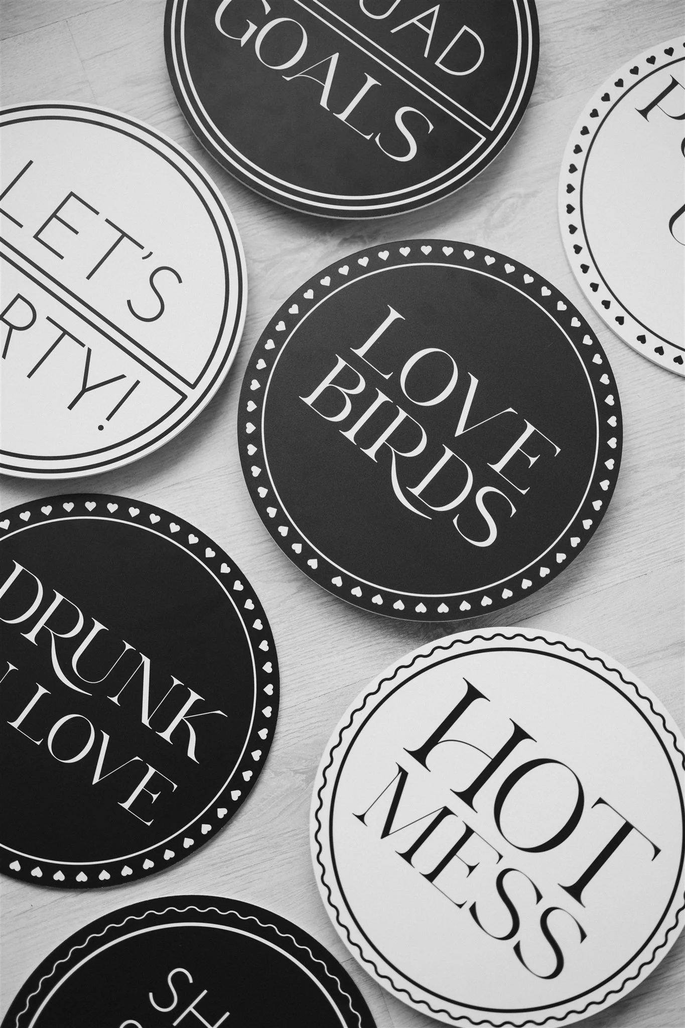 Black and white circular signs with messages like "Love BIrds," "Hot Mess," "Let’s Party," "Drink Love," and "Road Trip" on a light wooden surface.