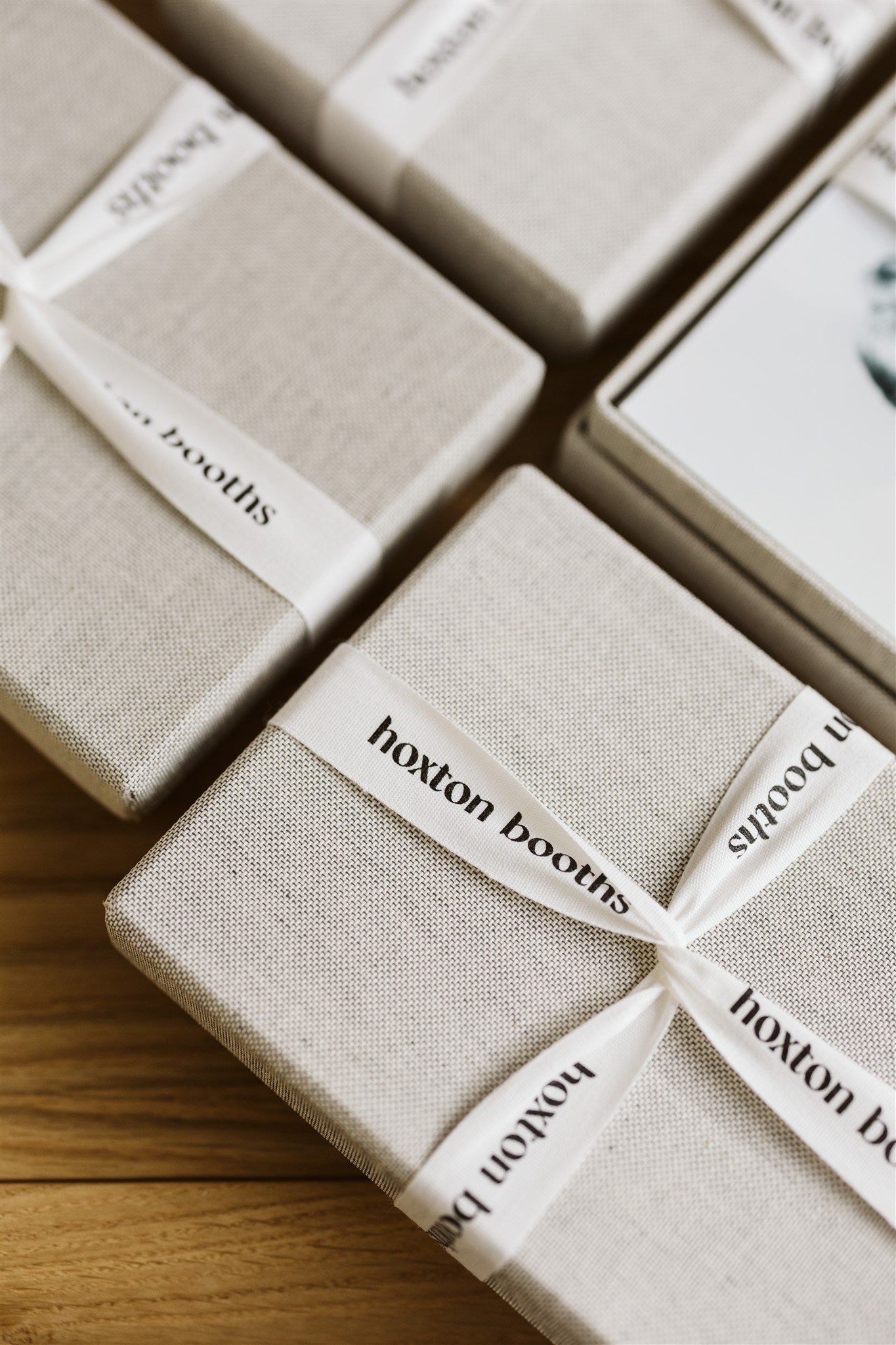 Stacked beige fabric gift boxes with white ribbon that says 'Hoxton Booths' on a wooden surface.