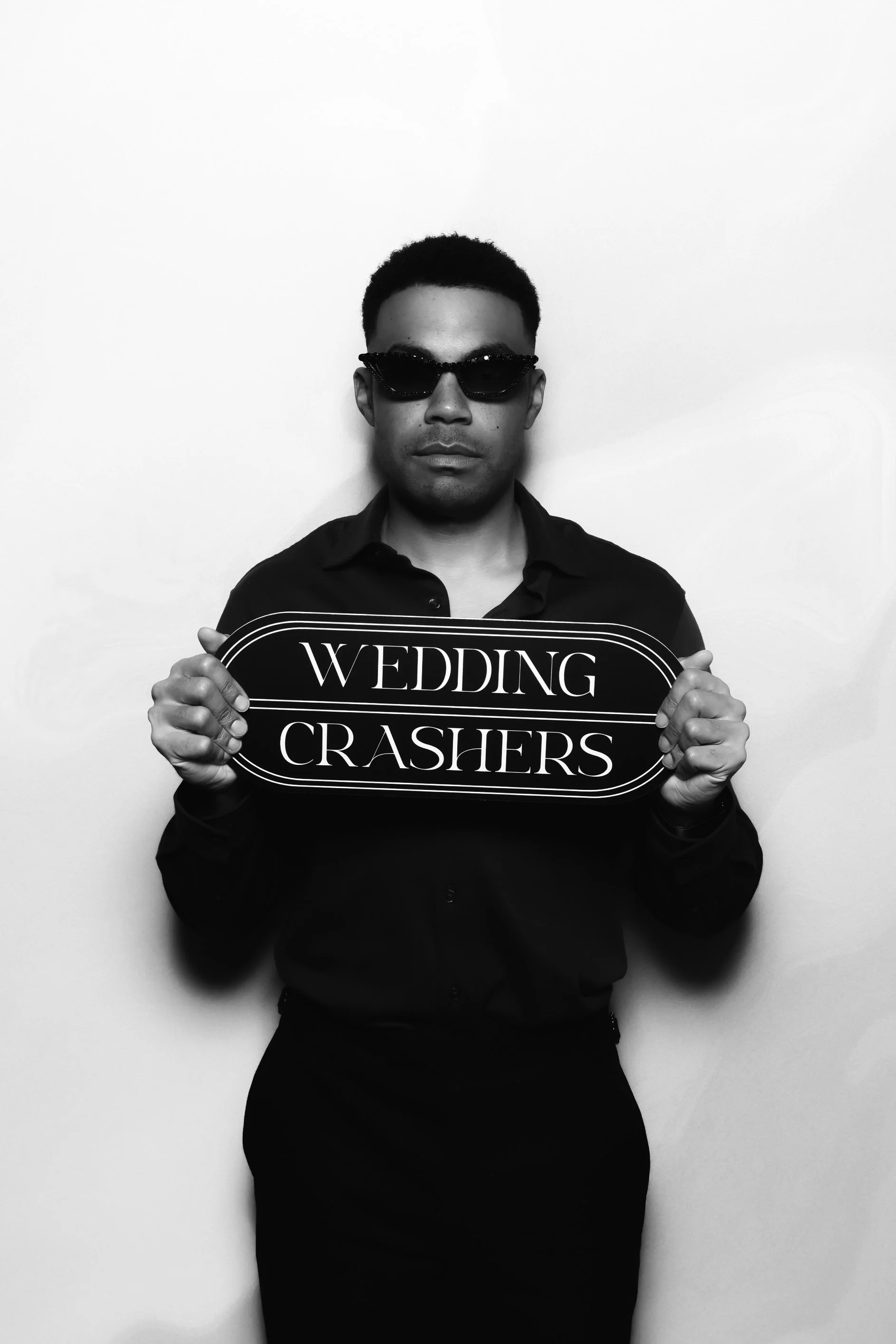 A man wearing dark sunglasses and a dark shirt holding a sign that reads 'WEDDING CRASHERS' in front of a plain white wall.
