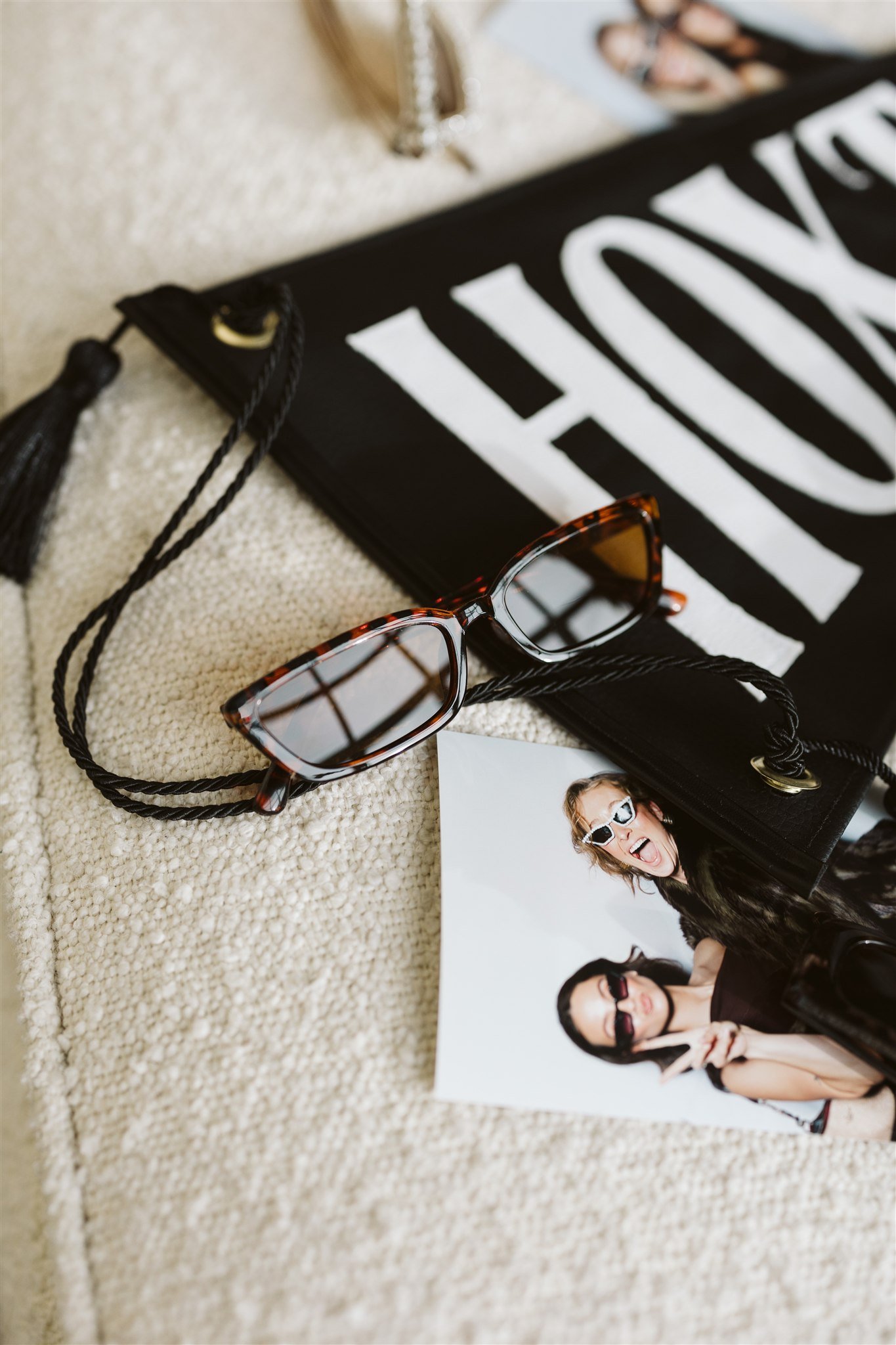 A pair of eyeglasses with tortoiseshell frames, a black handbag, a printed photo of two women wearing sunglasses, and part of a magazine with bold white letters on a black background, all arranged on a beige textured surface.
