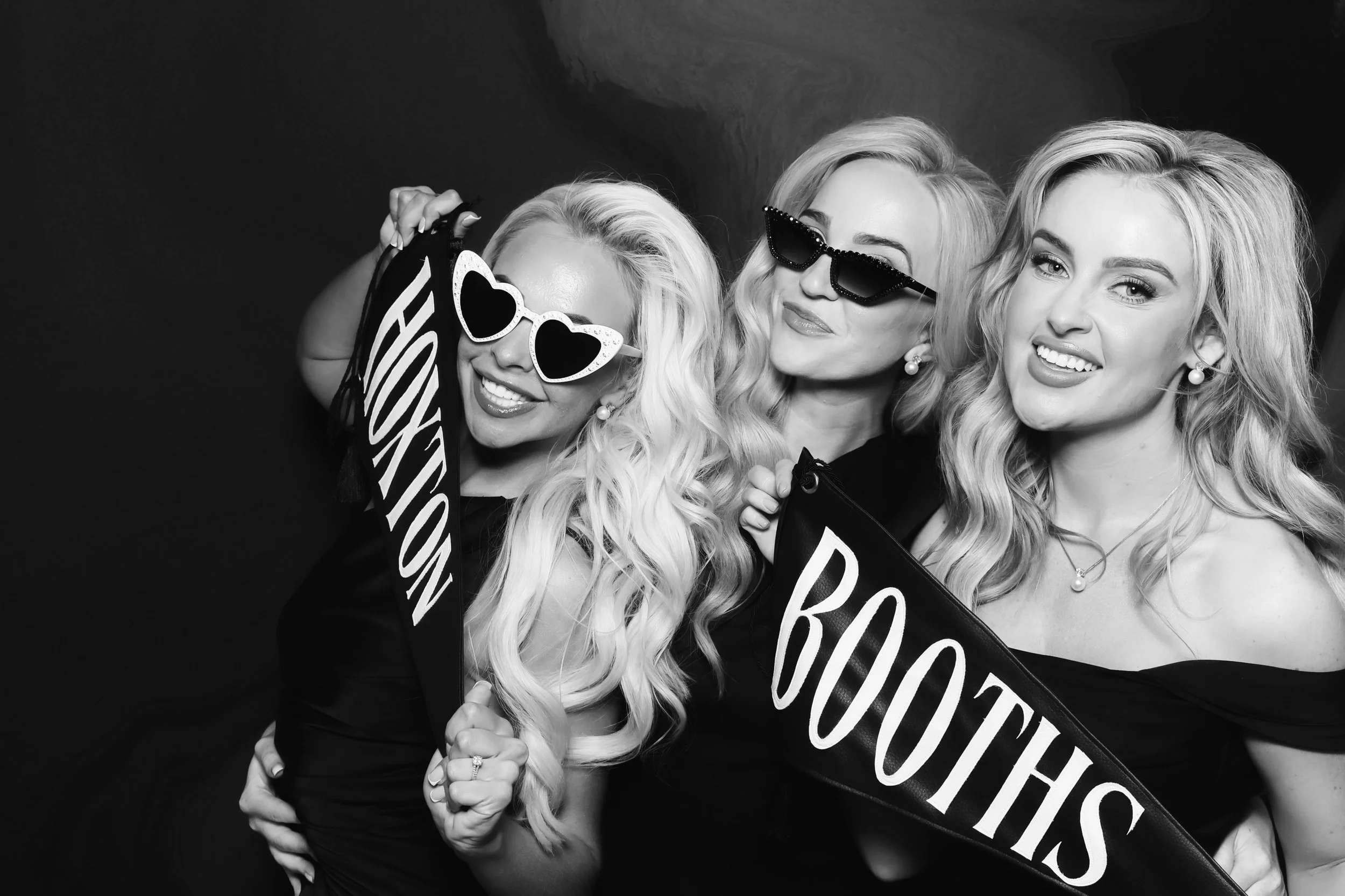Three women at a social event, wearing sunglasses and holding sign banners that read 'Hoxton' and 'Booths' smiling and posing for the camera.