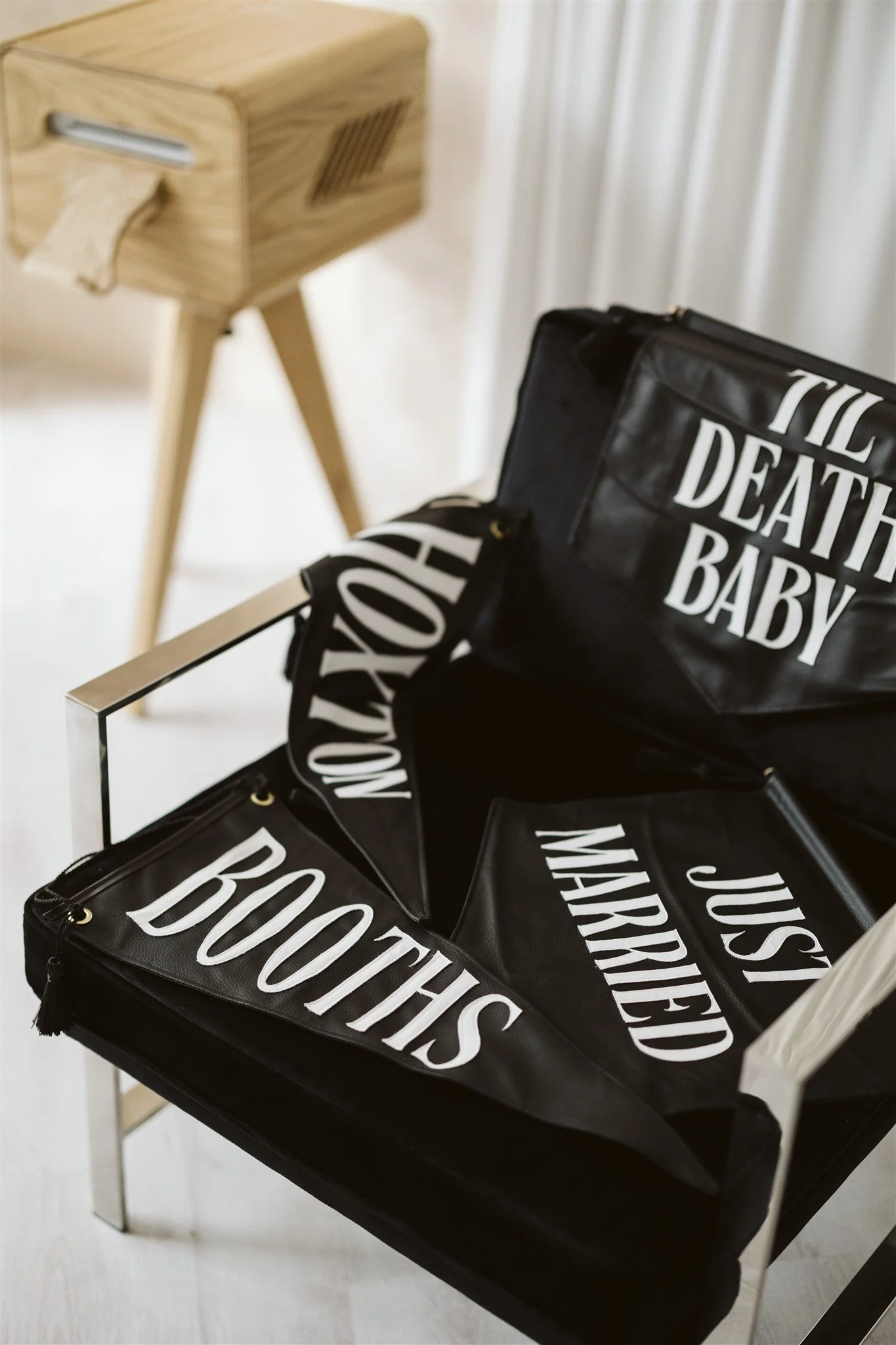 Black handmade leather flags with white text labeled 'JUST MARRIED' and a black bag with white text labeled 'BOOTHS' inside it, placed on a black chair.