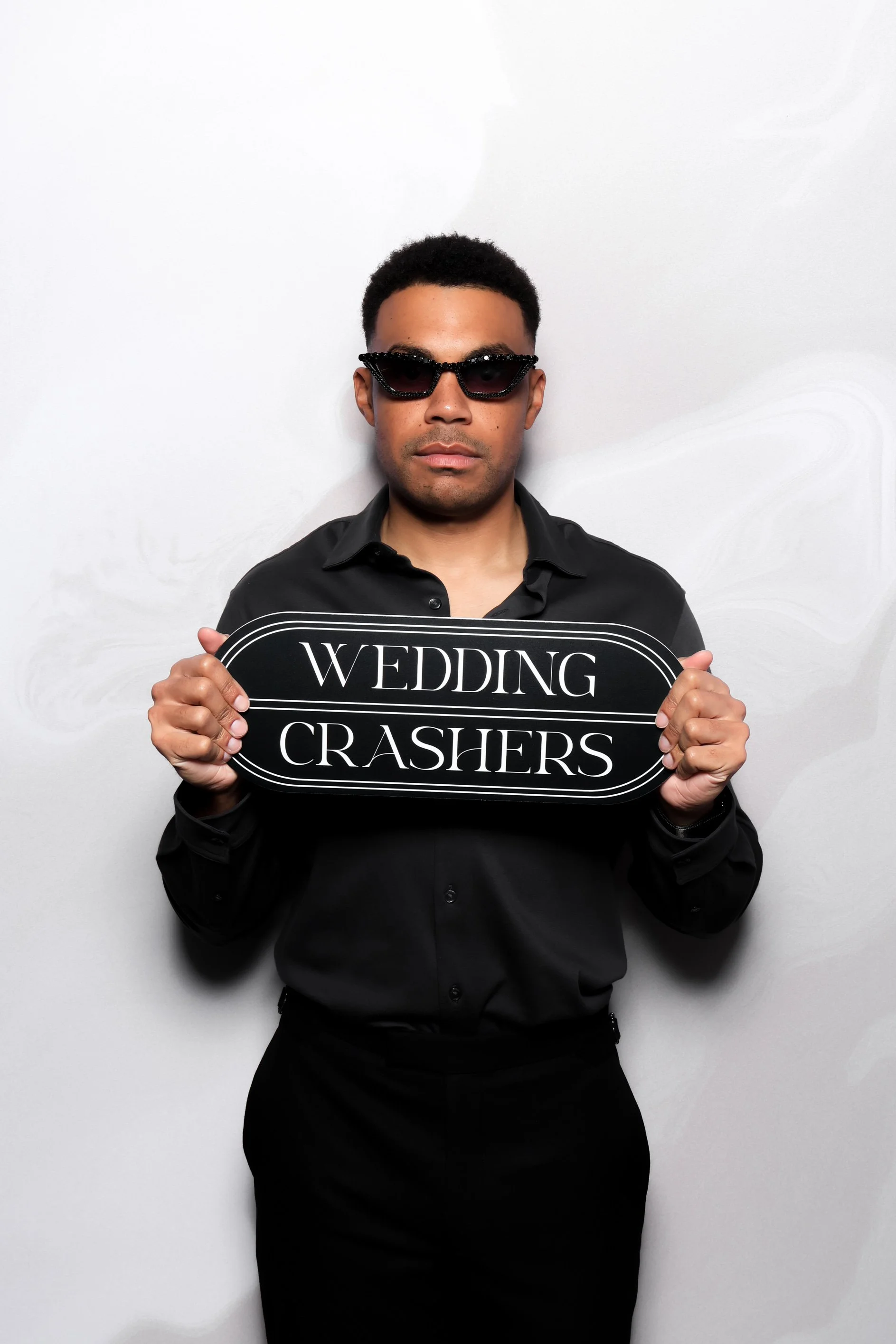 A man in black clothing and sunglasses holding a sign that reads 'Wedding Crashers'.