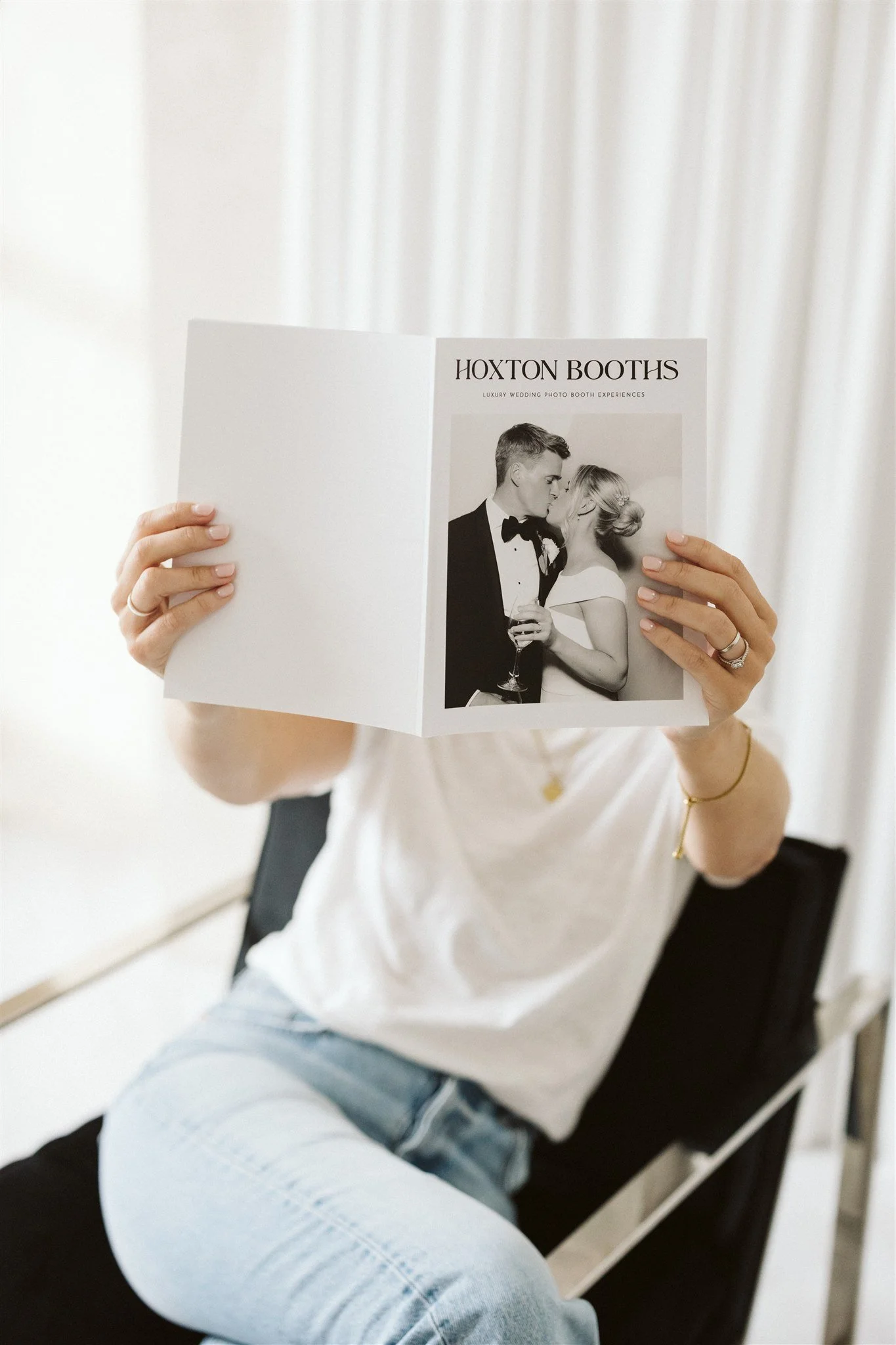 Person sitting on a black chair holding up a brochure with a wedding photo and the text 'Hoxton Booths' and 'Luxury Wedding Photo Booth Experiences'.