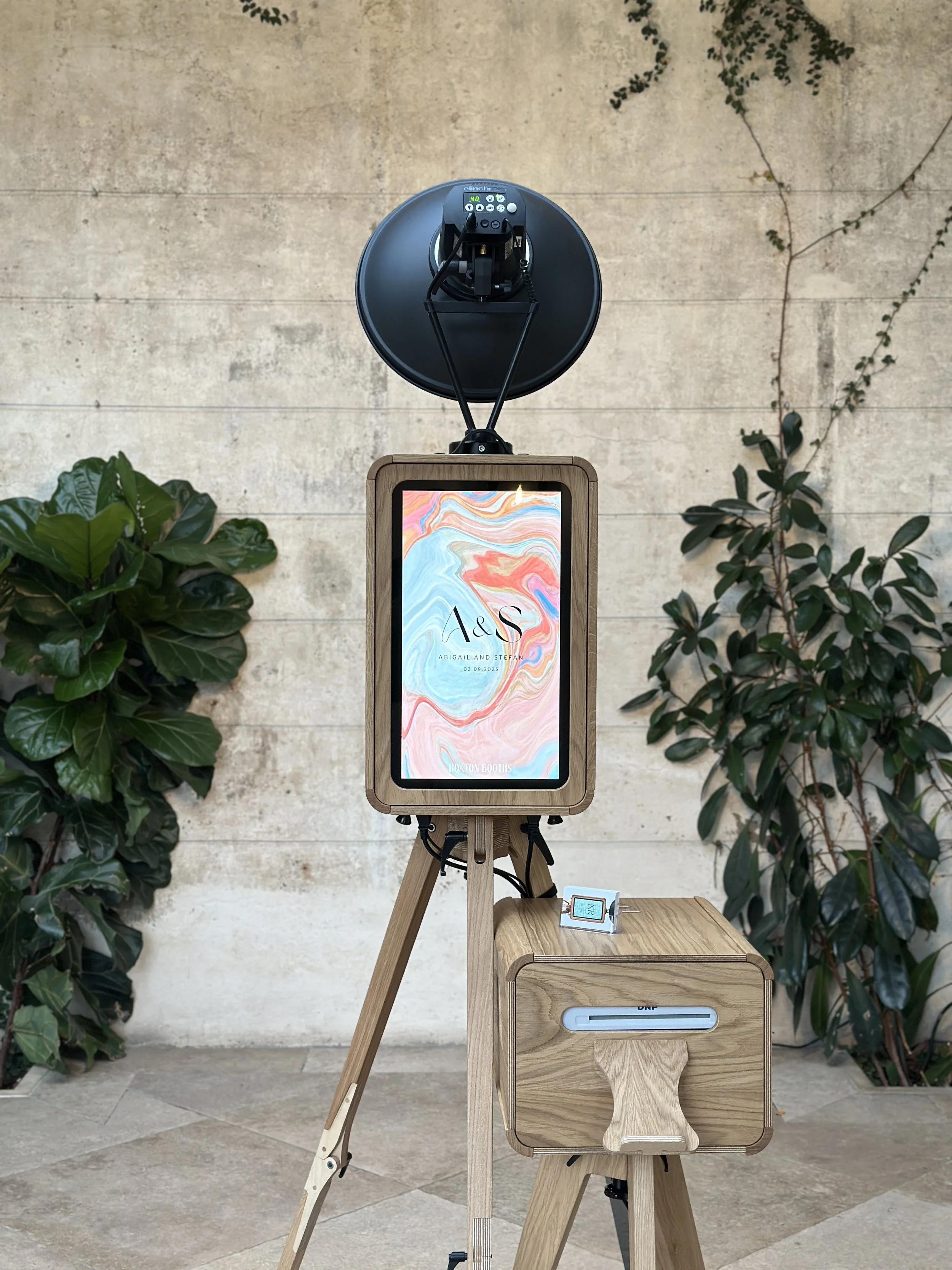 A photo booth setup featuring a wooden structure with a digital screen displaying decorative text, a black light mounted above, in front is the photo booths printer station, beautiful in wood, and greenery in the background.