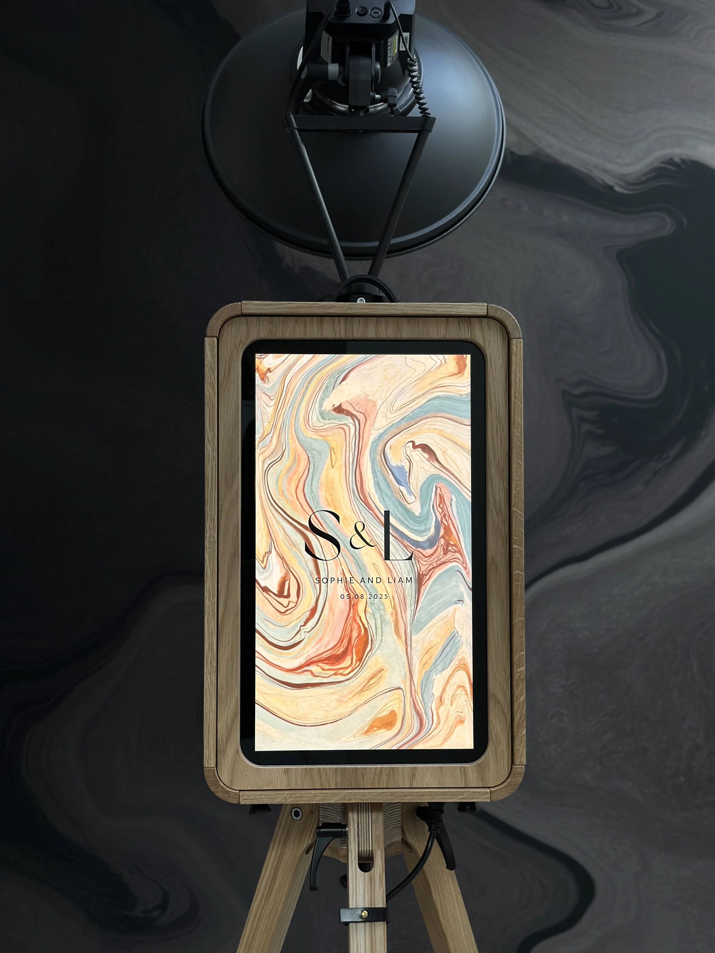 A digital display framed with wood, mounted on a tripod, showing a colorful abstract background with initials 'S & L' and text 'SOPHIE AND LIAM 05.08.2025'.