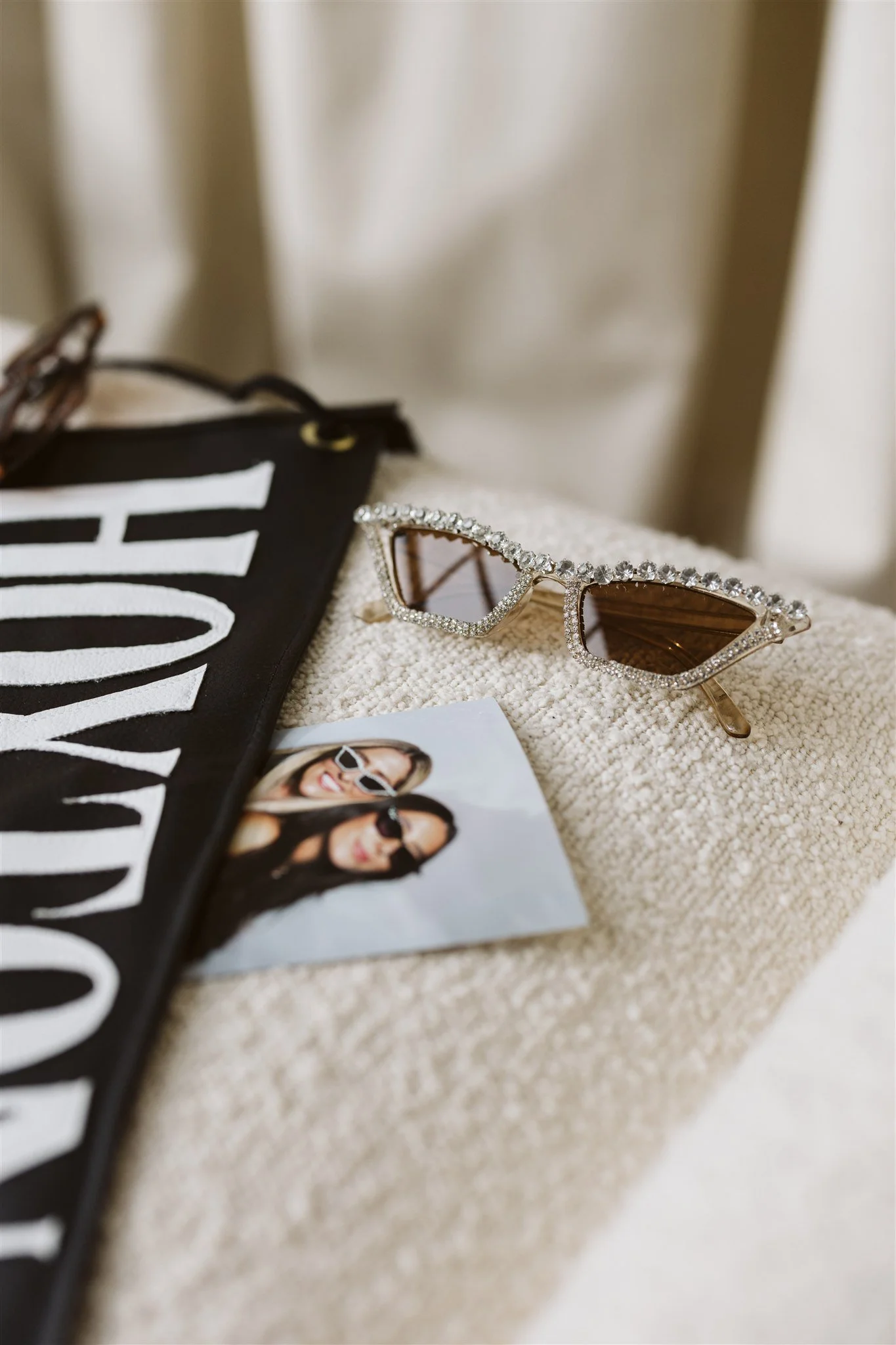 A pair of sunglasses with rhinestones on the frames resting on a textured off-white surface, along with a photograph of two women wearing sunglasses, and a black fabric item with white text.