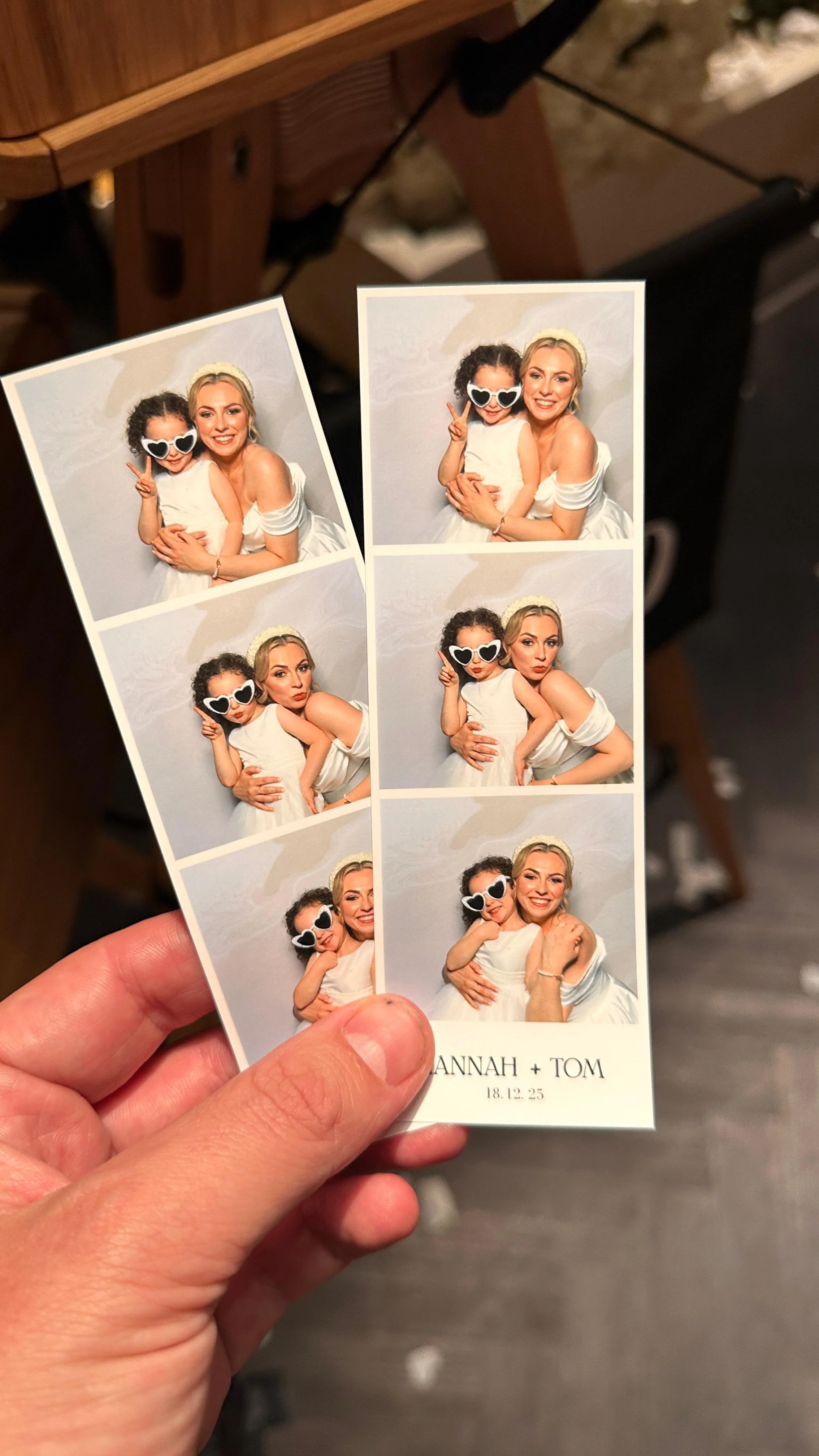 Photo booth strip with four pictures of two women, one with dark curly hair and one with blonde hair, wearing sunglasses and smiling, at a celebration event. Text at the bottom of the strip reads 'HANNAH + TOM' with the date '18.12.25'.