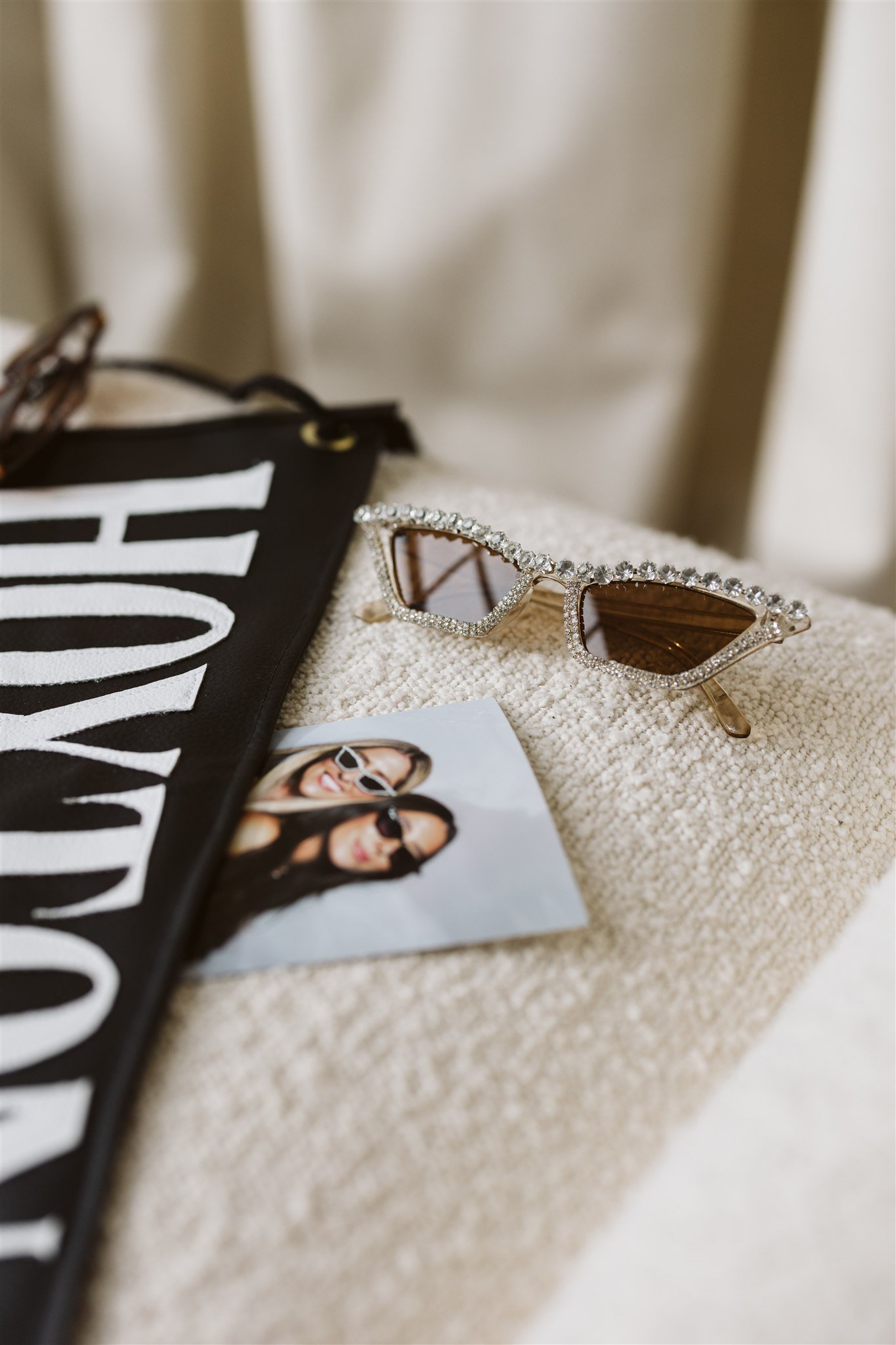 Pair of sunglasses with rhinestone-encrusted frames resting on a textured beige surface, partially covering a black fabric item with white text and a small photo of two women wearing sunglasses.