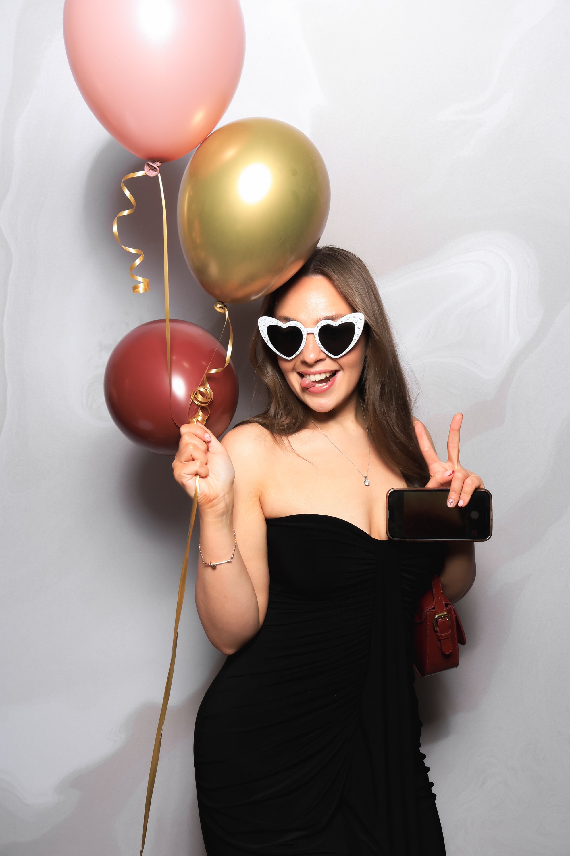 A woman in a black dress celebrating with pink, gold, and red balloons, wearing heart-shaped sunglasses, smiling, and holding a phone.