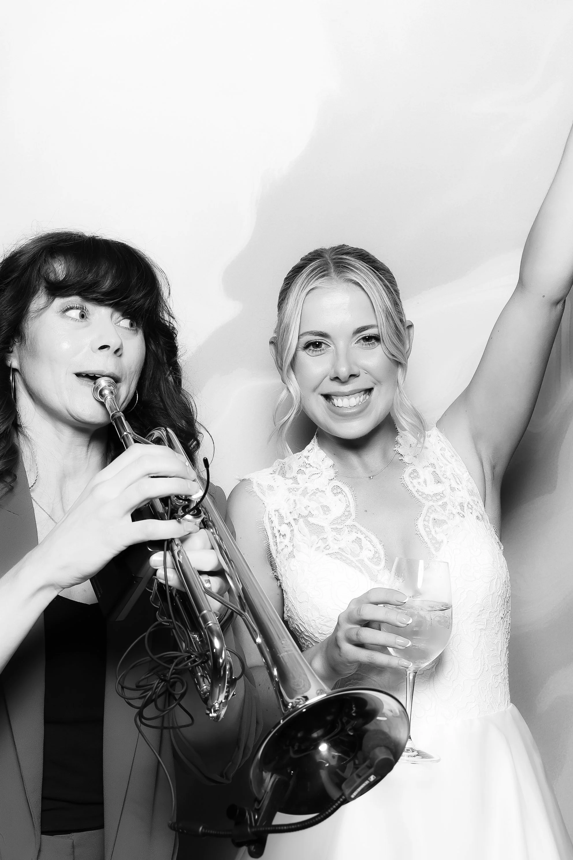 Black and white photo of two women enjoying a party; one is playing the saxophone and the other is smiling and holding a glass of white wine.