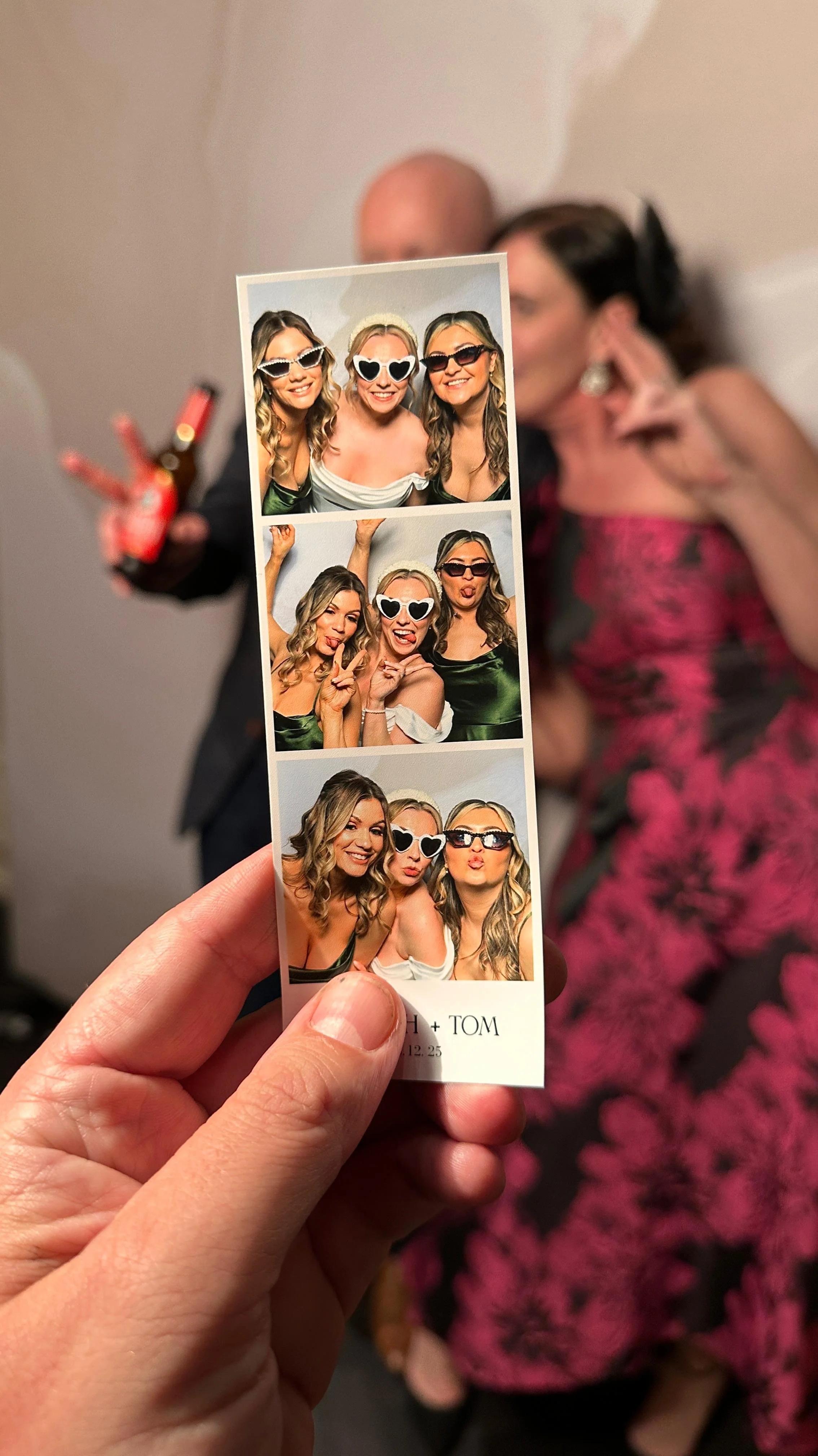 A strip of three photo booth pictures held by a hand, with three women wearing sunglasses making playful faces. In the background, there are two people, a woman in a pink and black dress and a man in a suit, slightly out of focus.