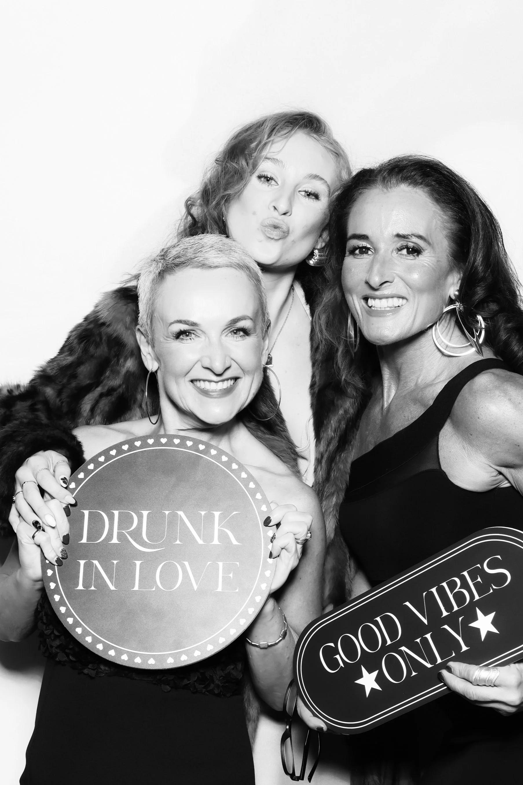 Three women at a party, smiling and holding signs that say "Drunk in Love" and "Good Vibes Only" in a black-and-white photo.