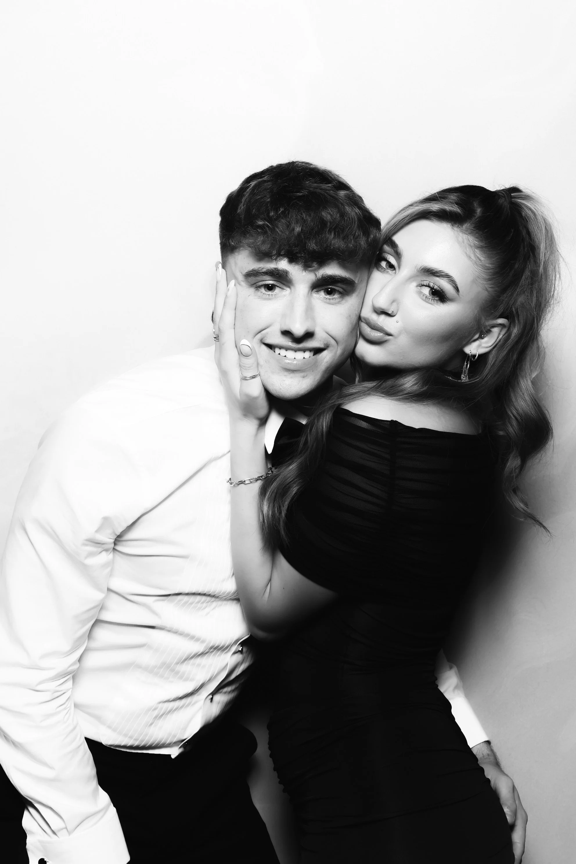 A black-and-white photo of a young man in a white shirt and young woman in a black dress posing closely together against a plain background, with the woman's arm around the man's shoulder and their faces next to each other.