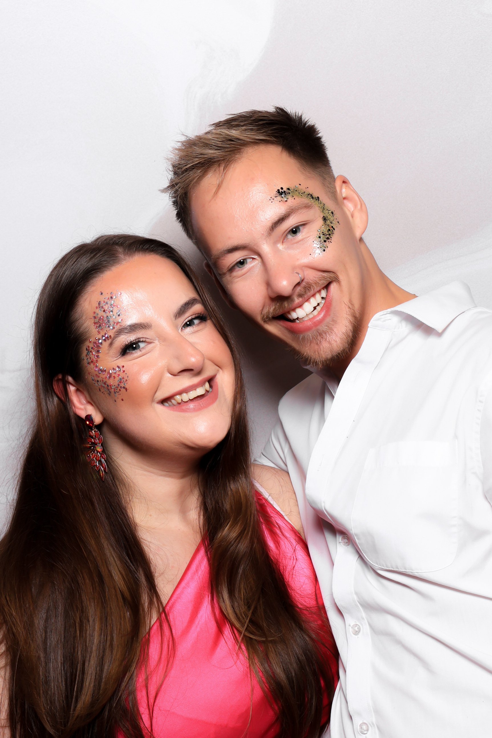 Two friends smiling for a selfie with glitter on their faces, one wearing a pink dress and the other in a white shirt, against a white background.
