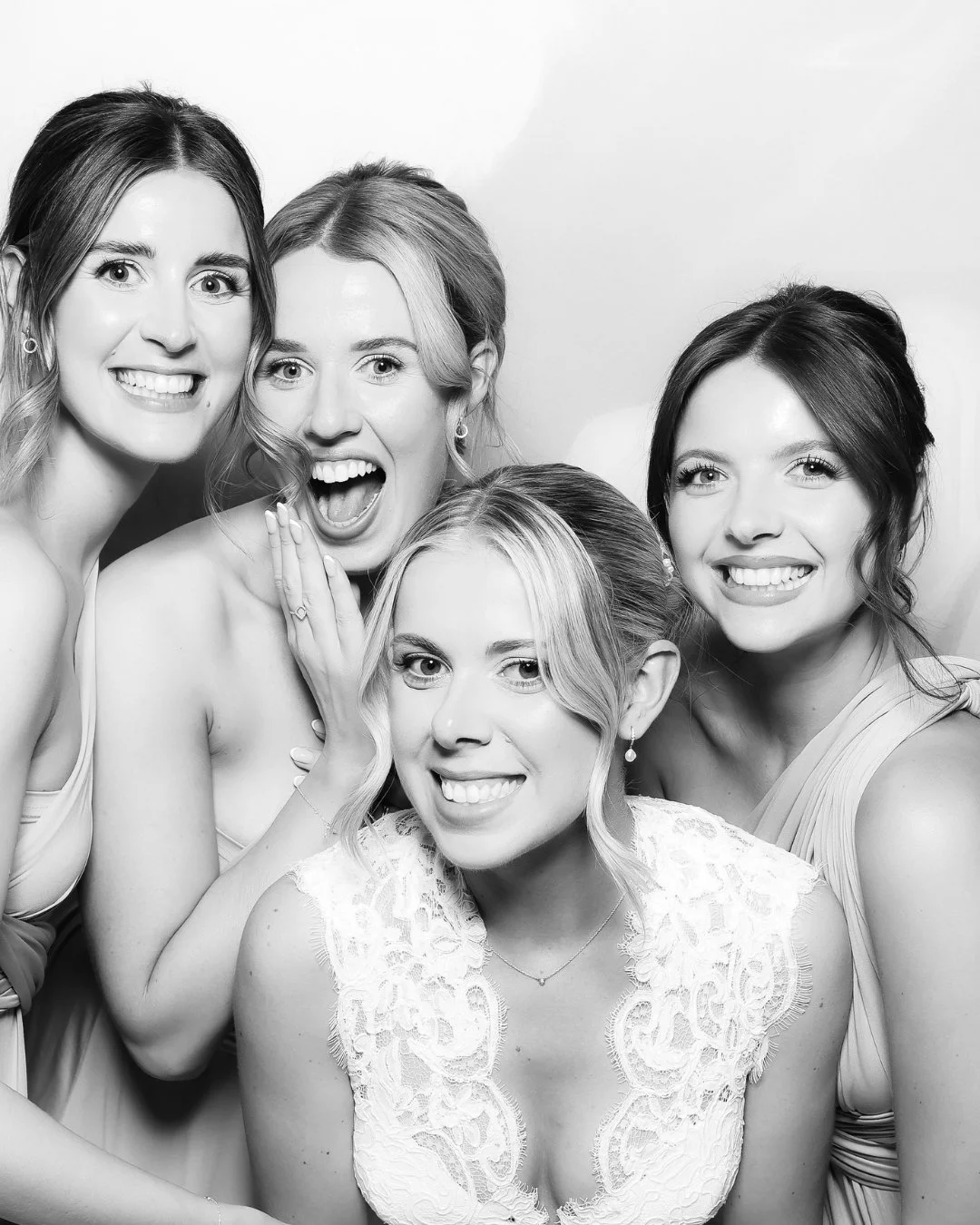 Black and white photo of five women smiling and posing together, dressed elegantly, with one woman in the center wearing a lace dress.