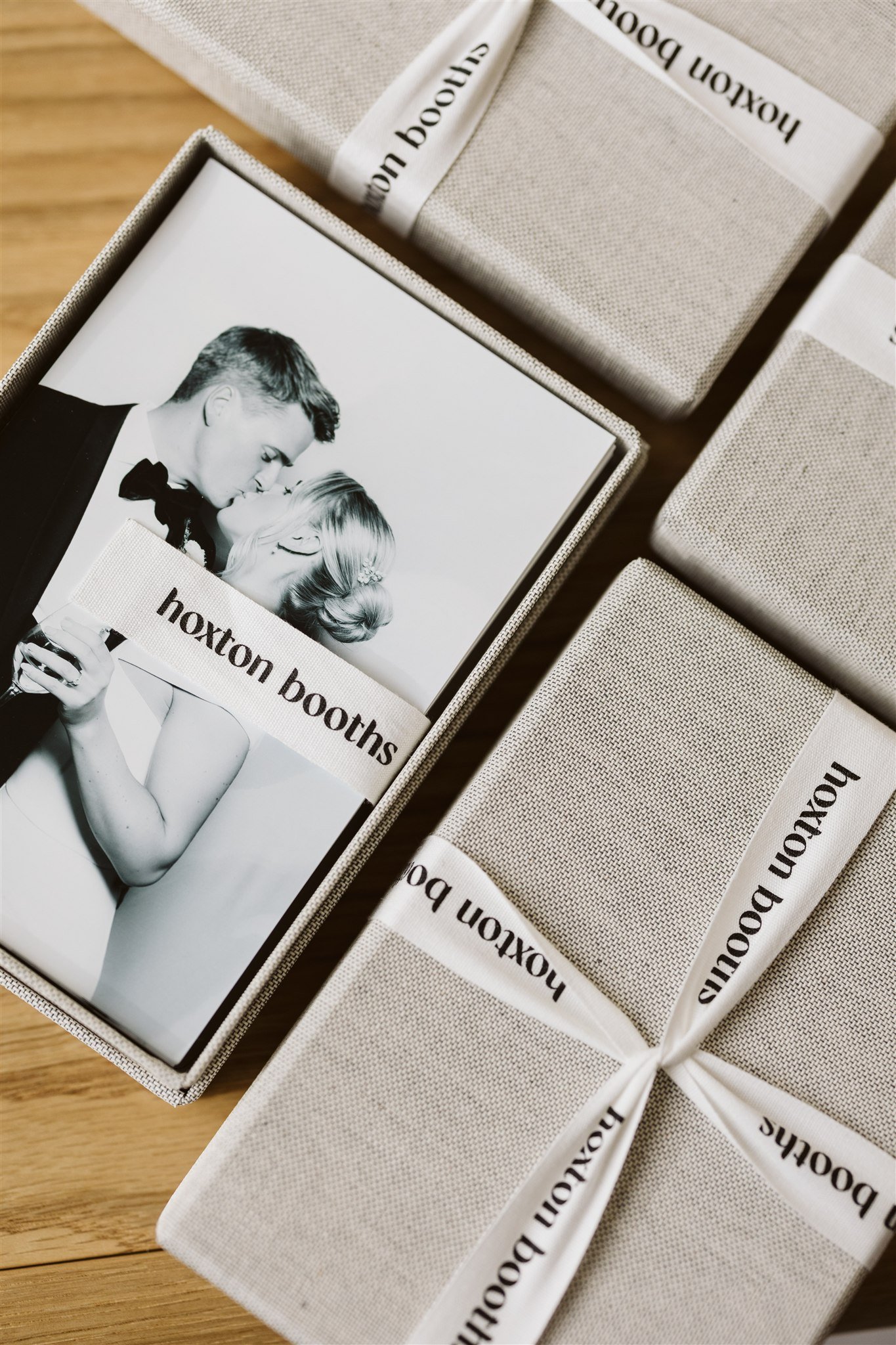 A black-and-white photograph of a couple dressed in formal attire, kissing, inside a box labeled 'Hoxton Booths', surrounded by wrapped gift boxes on a wooden surface.
