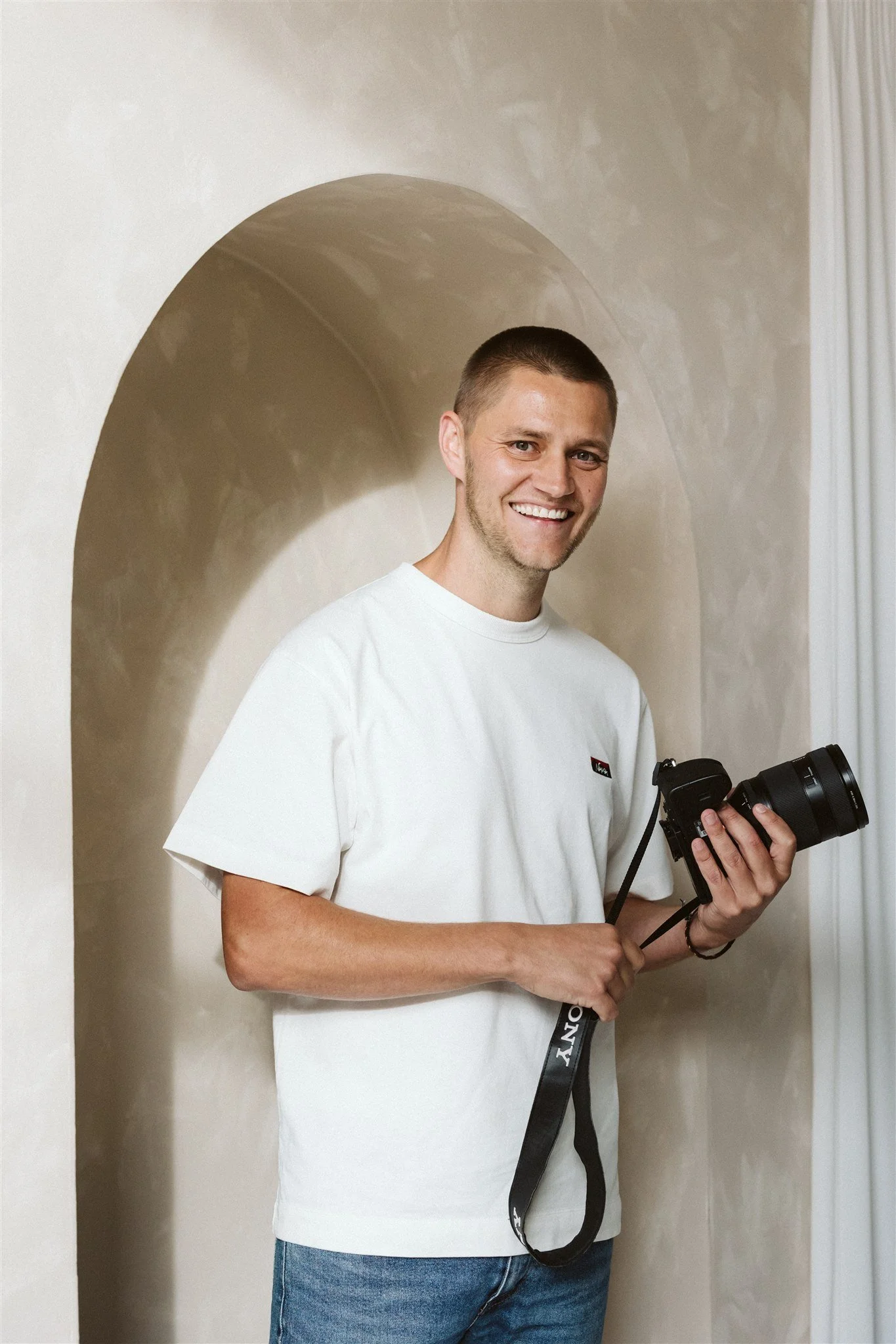 A smiling man holding a DSLR camera indoors near a beige wall with an arched alcove.