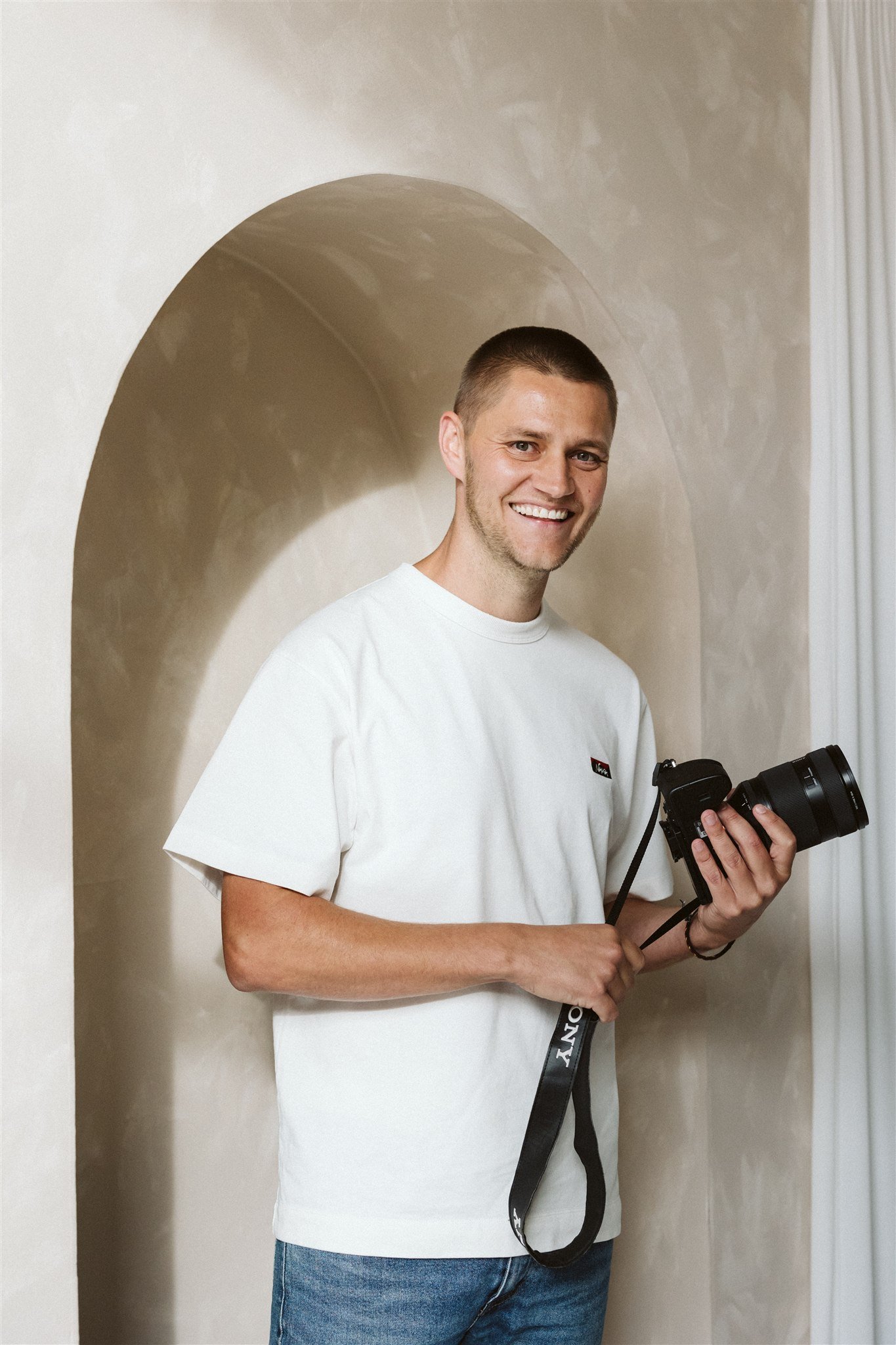 A smiling man in a white t-shirt holding a camera, standing in front of an arched wall.