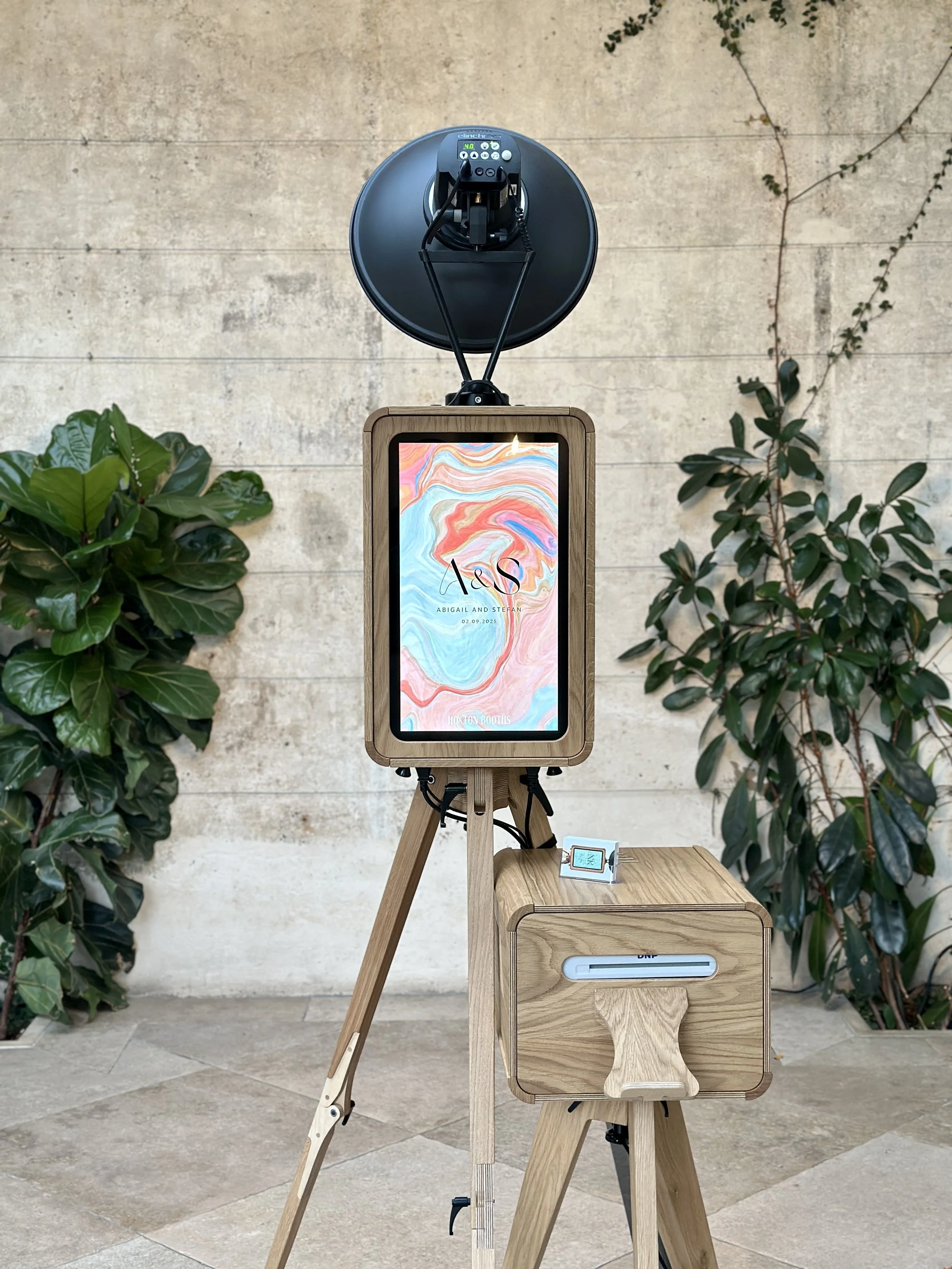 A photo of a photo booth setup with a digital screen, camera, and lighting equipment. The display shows an abstract background with text "A & S" and the names Abigail and Stefan, dated 02.09.2023. The setup is against a stone wall with green plants o