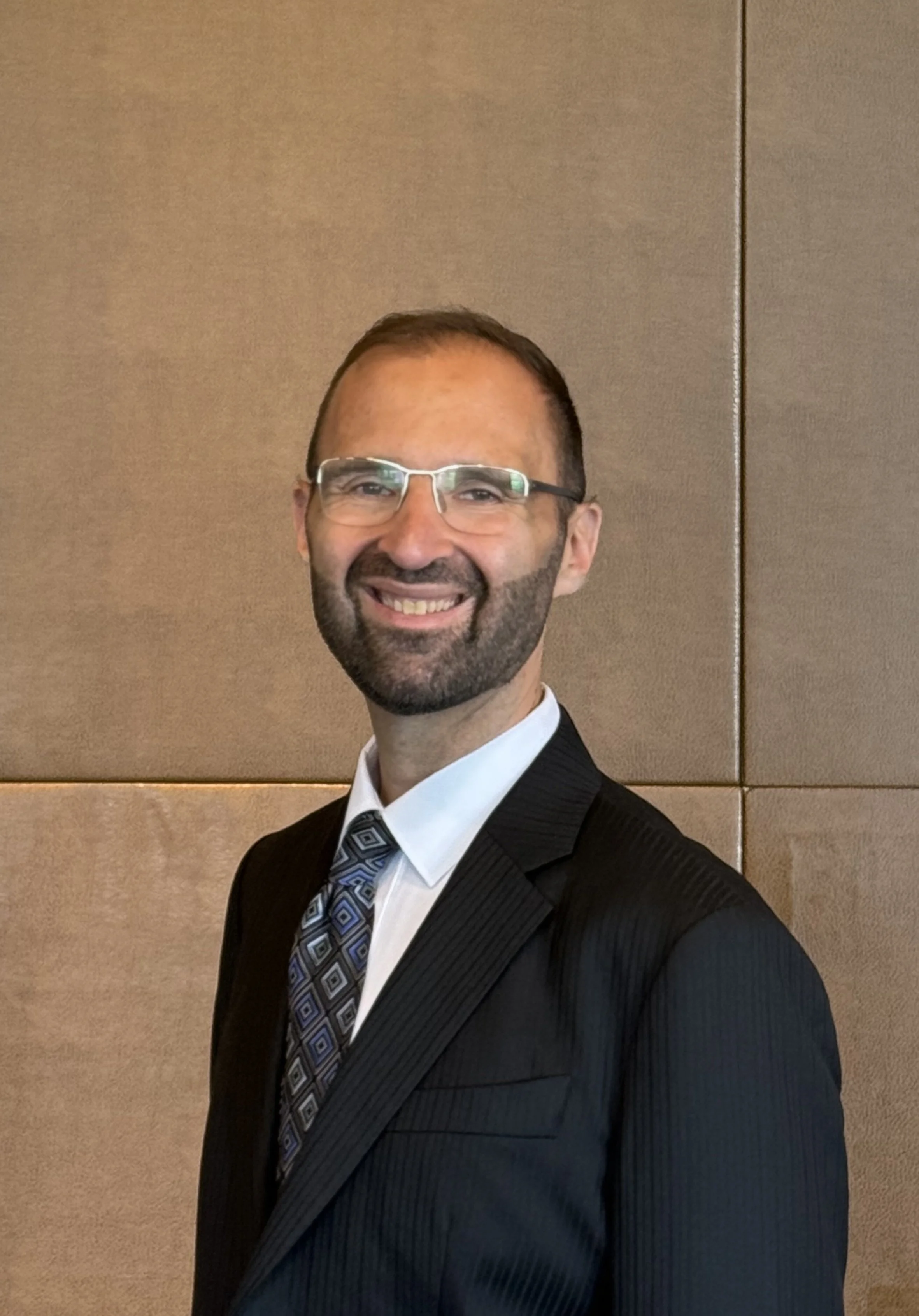 A smiling man in a suit and tie, wearing glasses, standing in front of a beige wall.