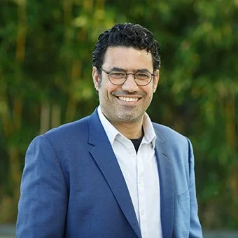 Smiling man in a blue suit and glasses outdoors with trees in the background.