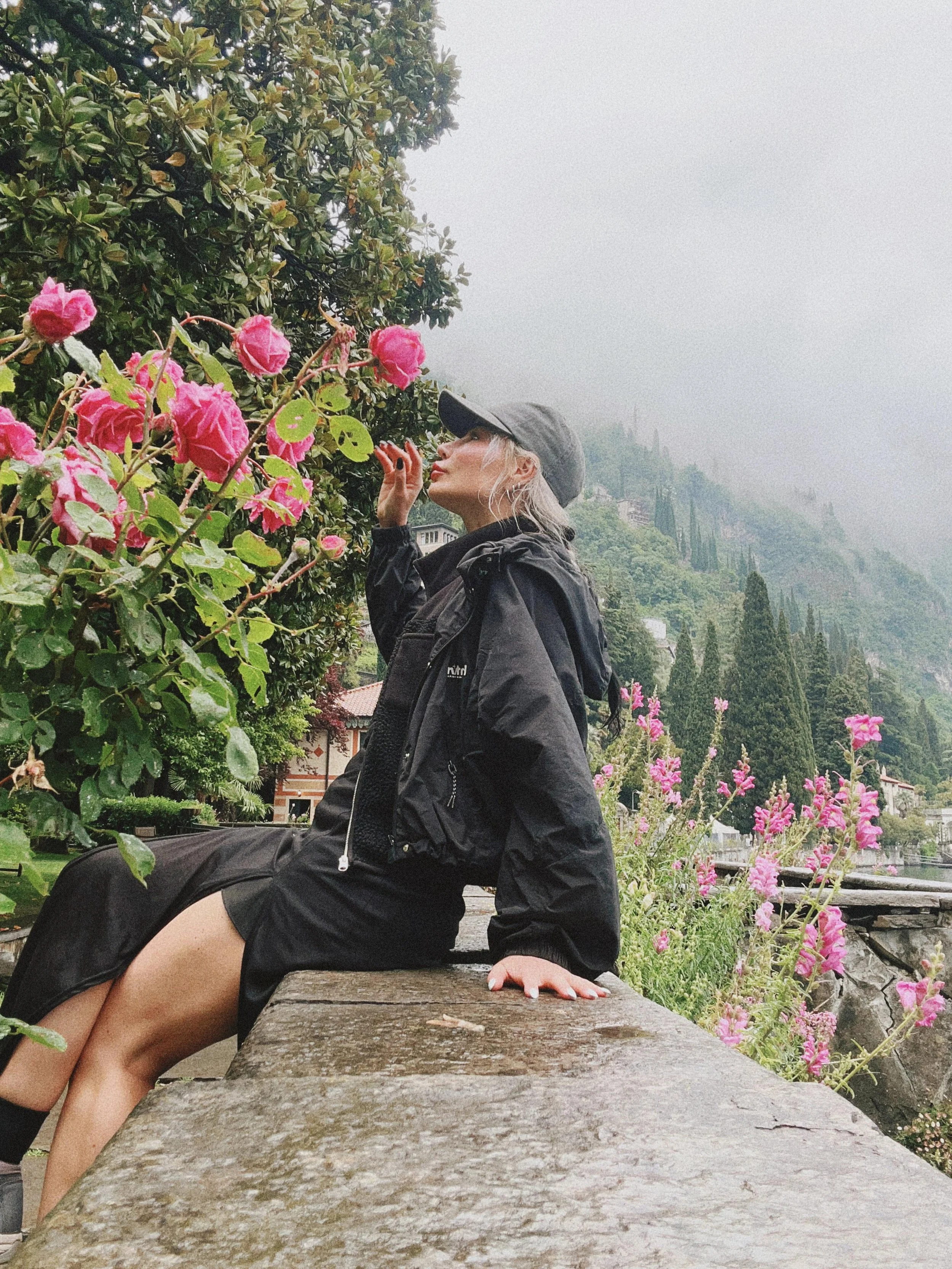 A woman with blonde hair wearing a black jacket, gray cap, and black dress sitting on a stone wall near pink flowers and greenery, with mountains and fog in the background.