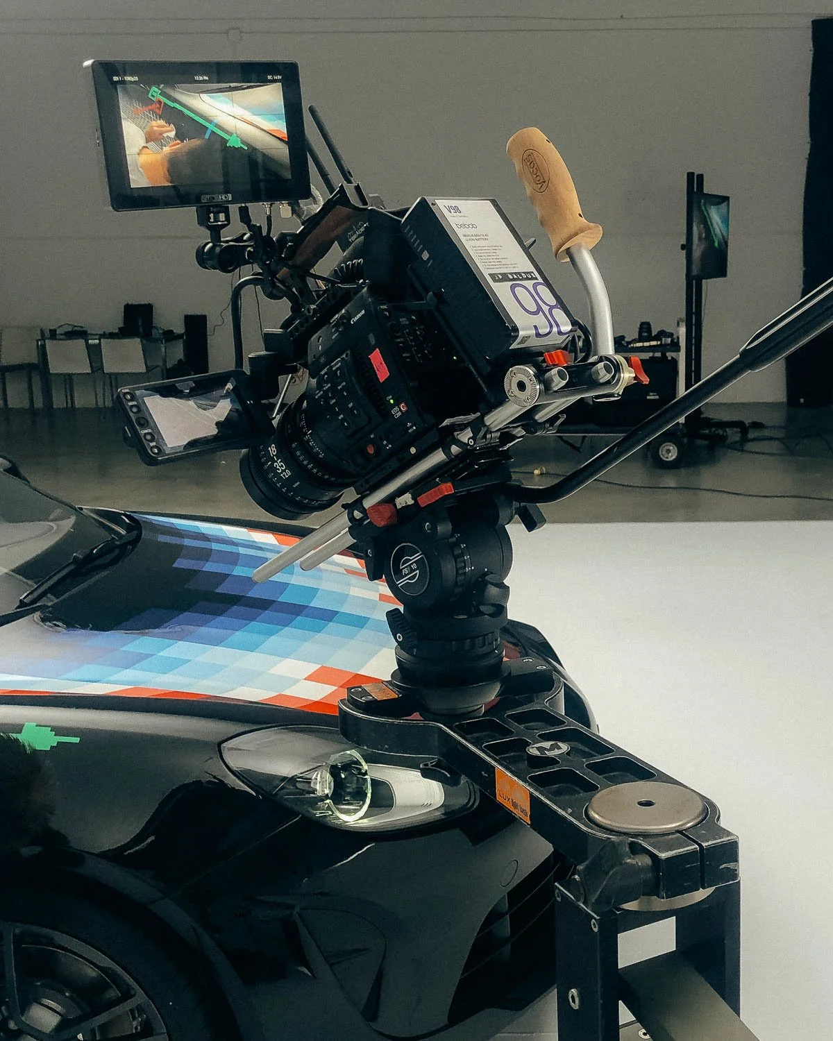 Close-up of a professional camera mounted on a car, likely on a racing circuit, with a monitor displaying a live view of a driver in a yellow suit on the racetrack.