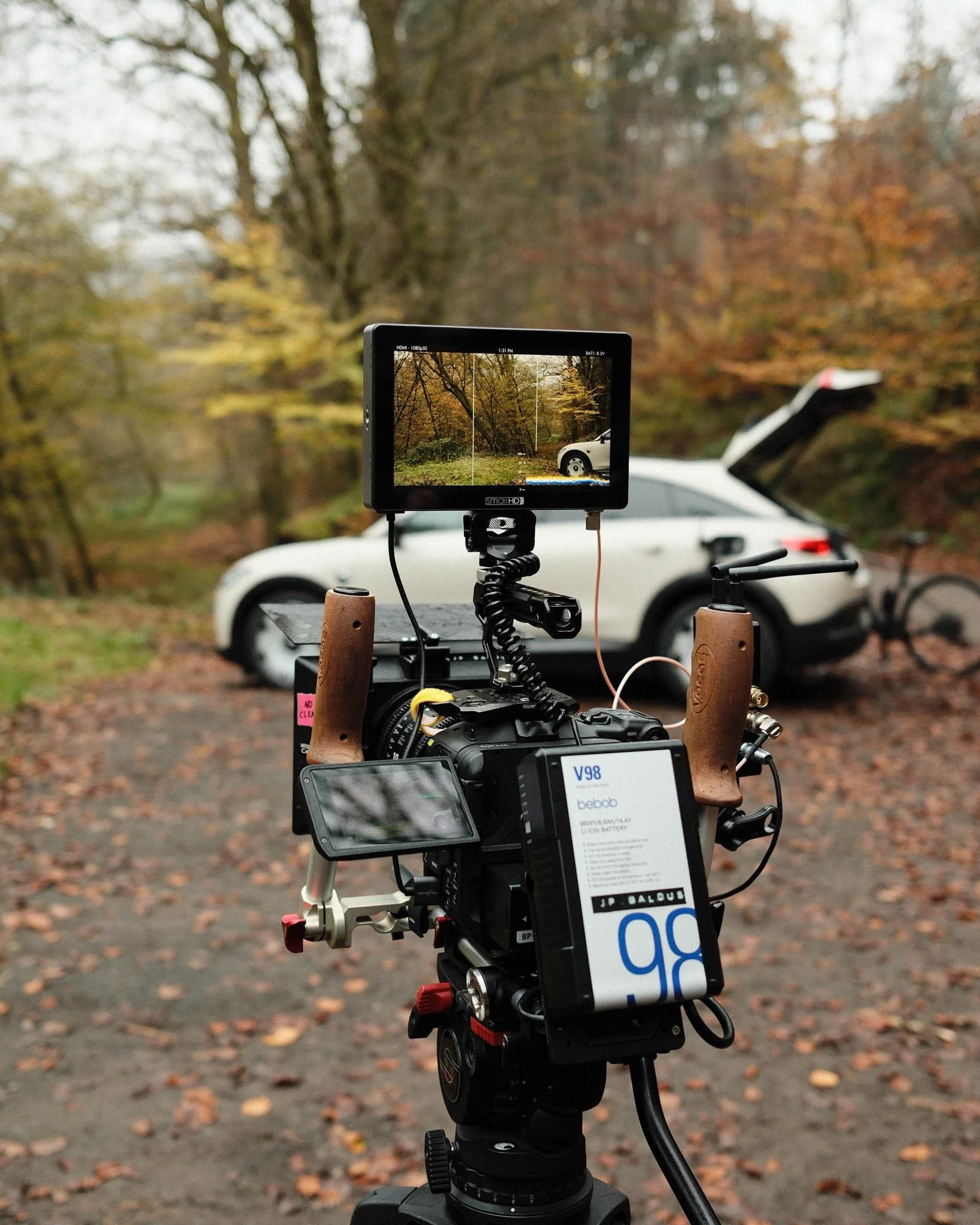 A camera on a tripod capturing footage of a white car with its trunk open, parked on a dirt ground surrounded by trees with autumn leaves.