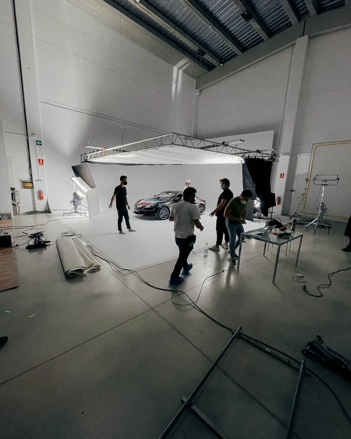A car photoshoot setup in a warehouse with a black sports car at the center, surrounded by crew members adjusting equipment, lights, and backdrop, with studio equipment and tools visible around the space.