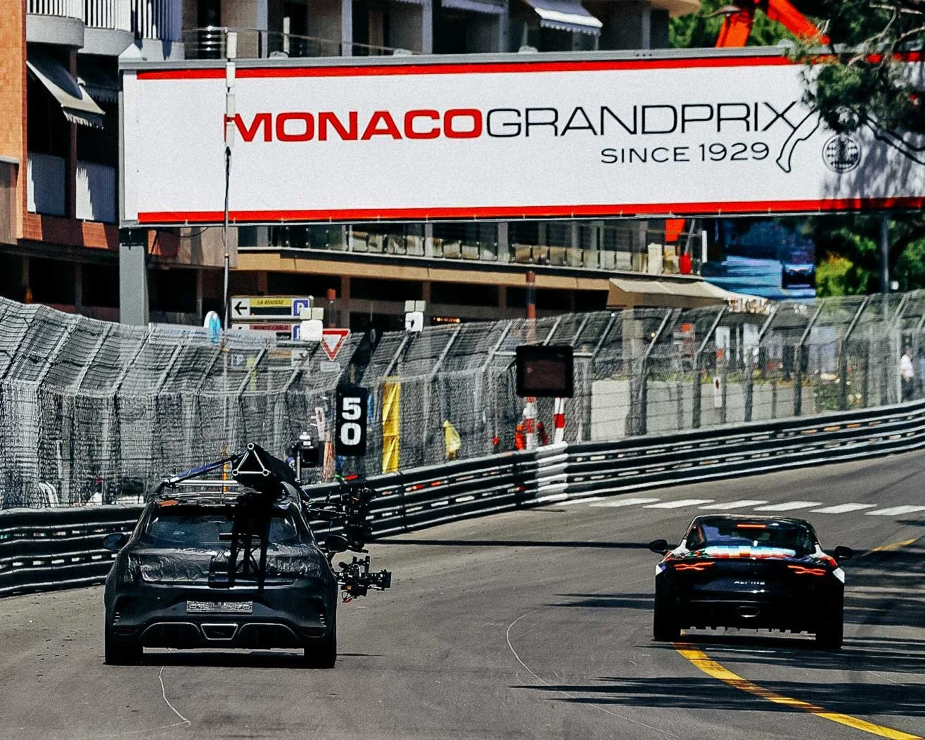 Two racing cars on a track, one camera car with equipment behind, and a Monaco Grand Prix banner overhead.