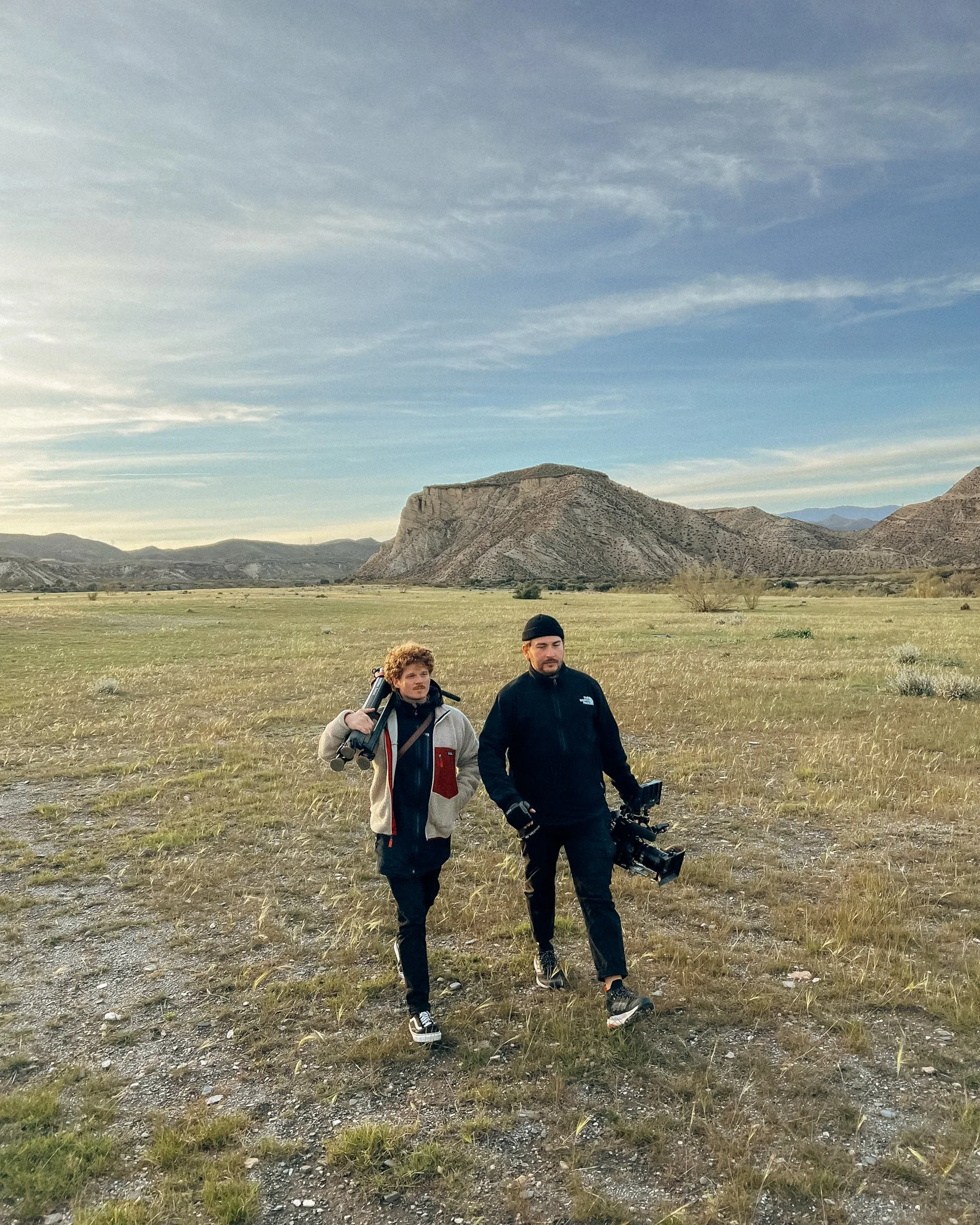Two men walking through a grassy field with mountains in the background. One man is carrying a tripod, and the other is holding a video camera.