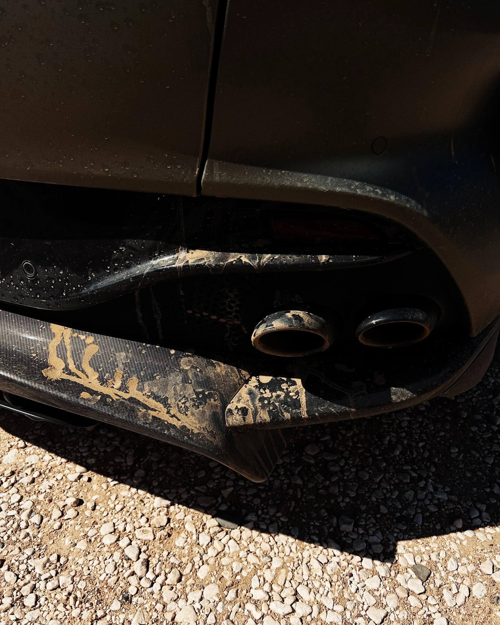 Close-up of the rear end of a dirty black car showing dual exhaust pipes and mud splatters.